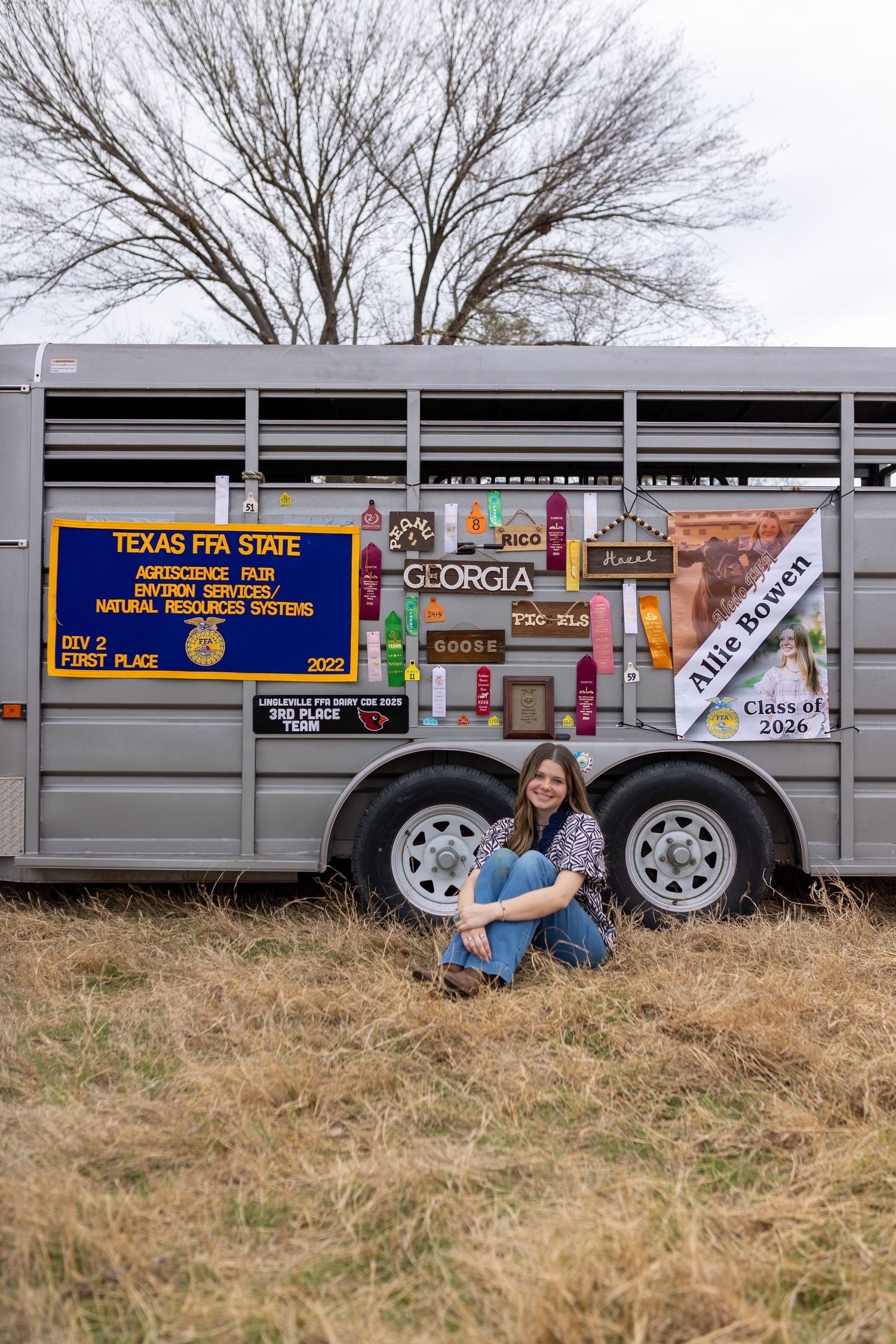 A livestock trailer decorated with various stickers and banners parked on grass with bare trees in the background.