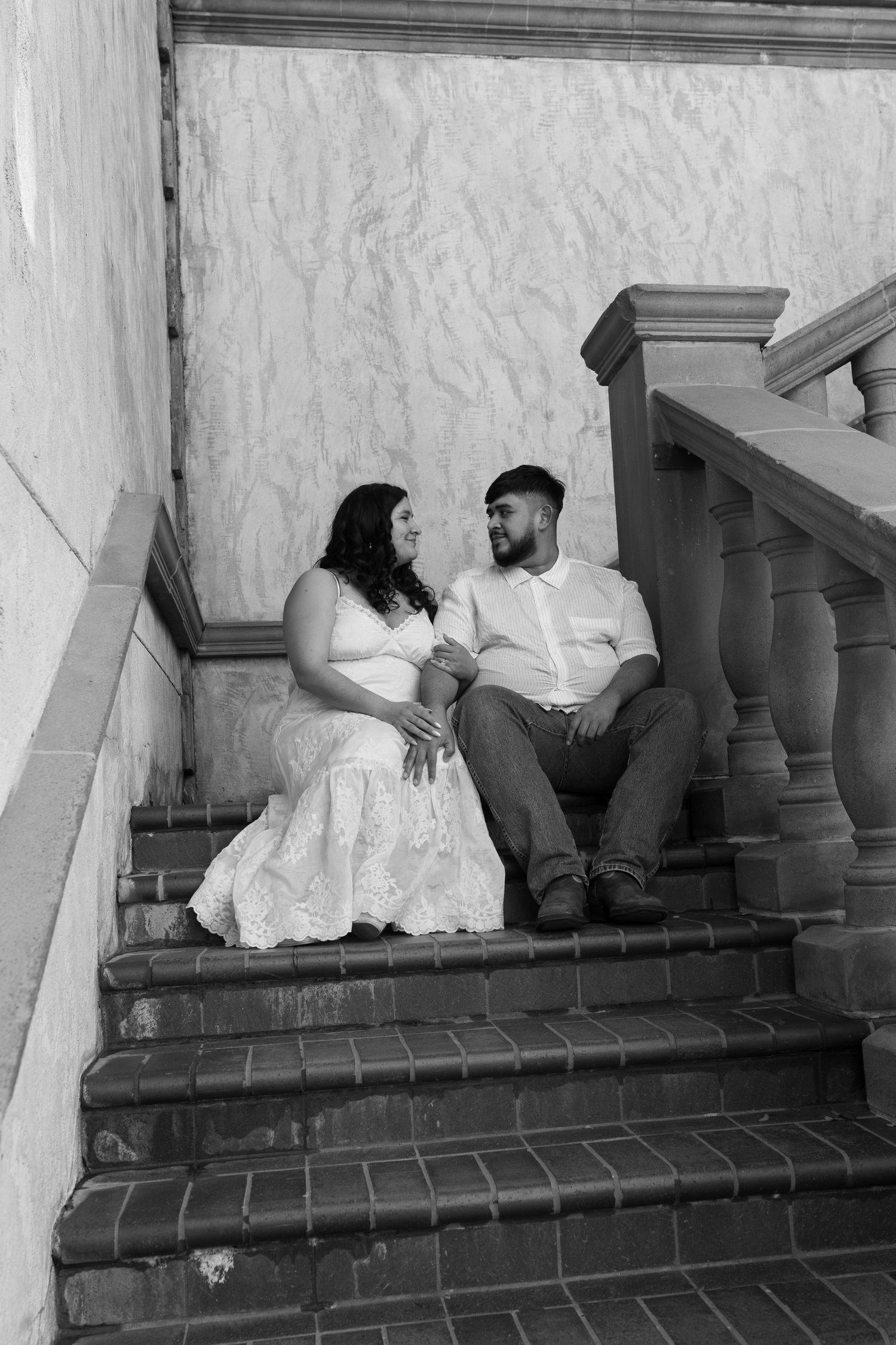 Black and white photo of a couple sitting on brick stairs, gazing at each other, woman in white lace dress, man in white shirt.