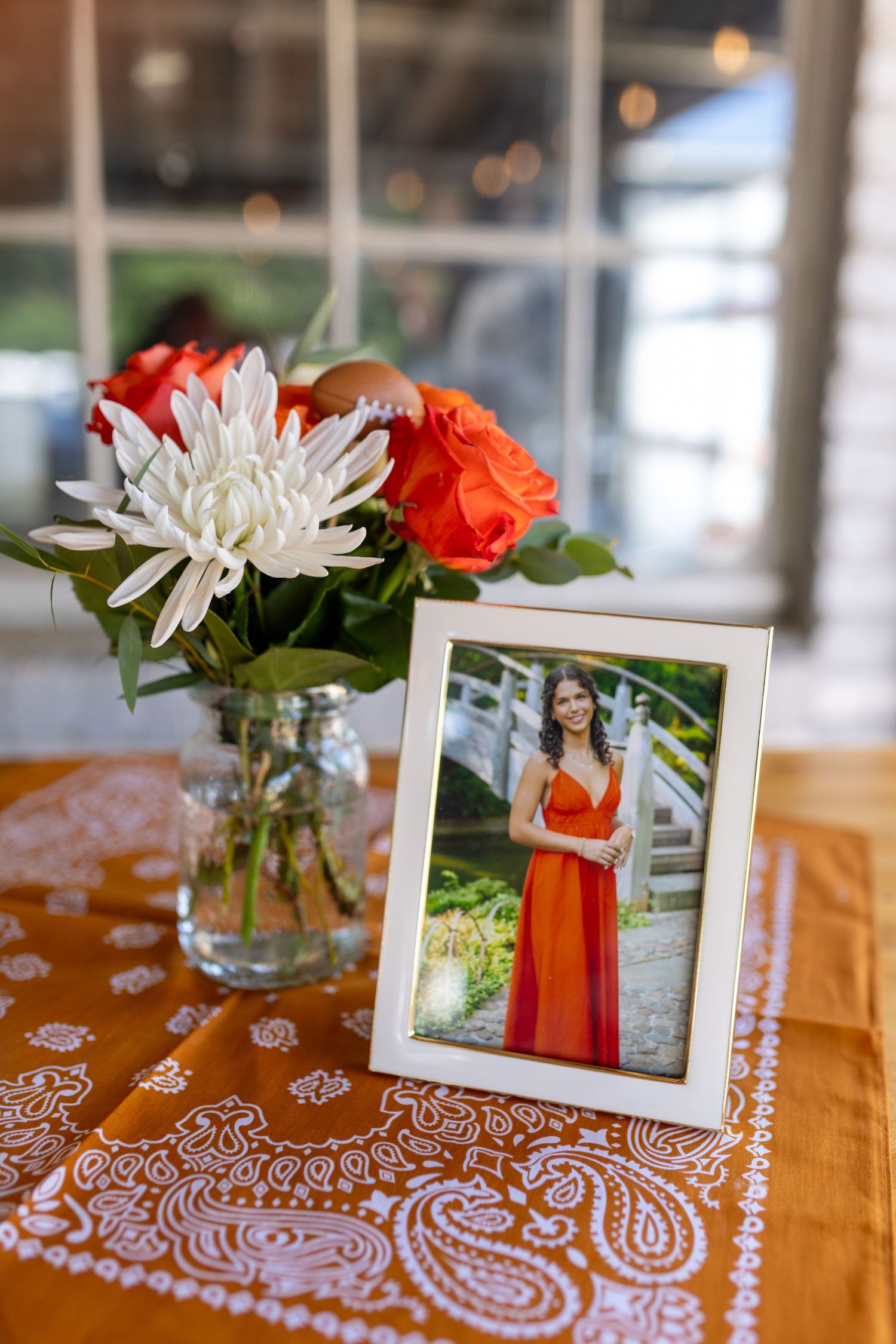 Framed photo of a woman in an orange dress displayed on a bandana tablecloth beside a mason jar with roses and chrysanthemums.