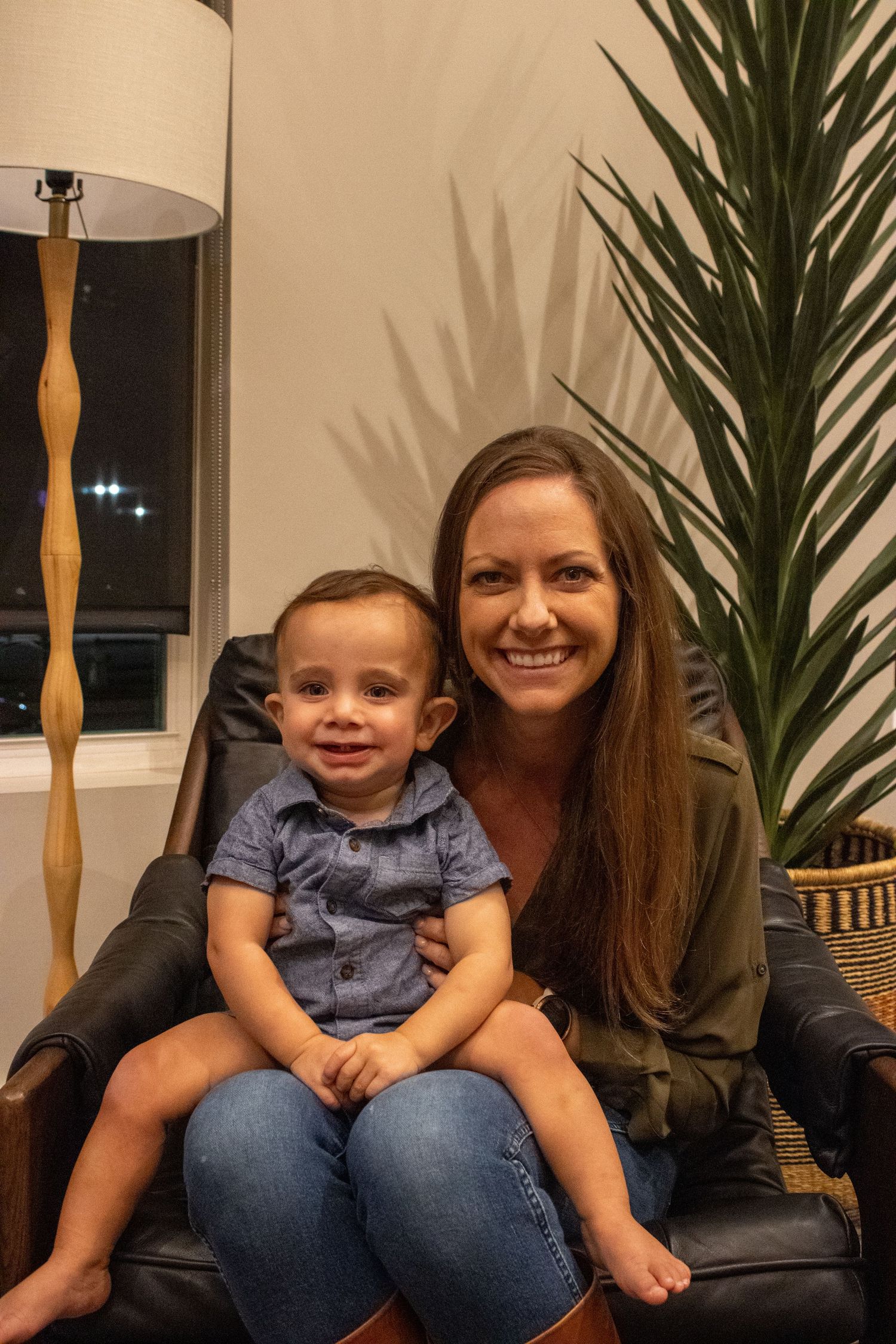 A casual indoor portrait on a leather chair with a decorative palm plant and modern lamp in the background.
