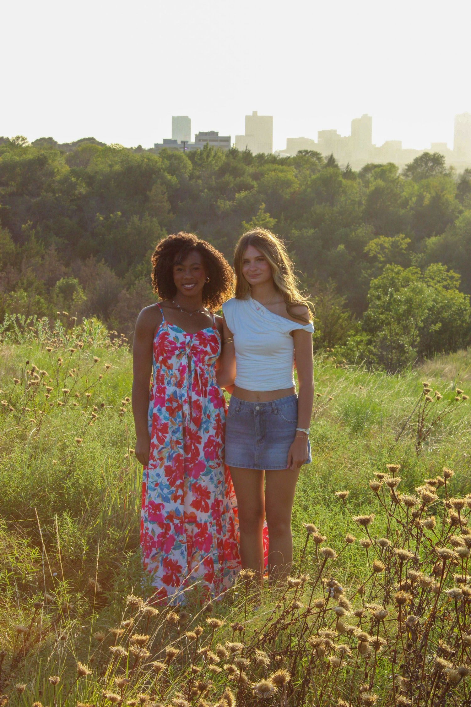 Friends posing in a sunlit meadow with city skyline visible in background.