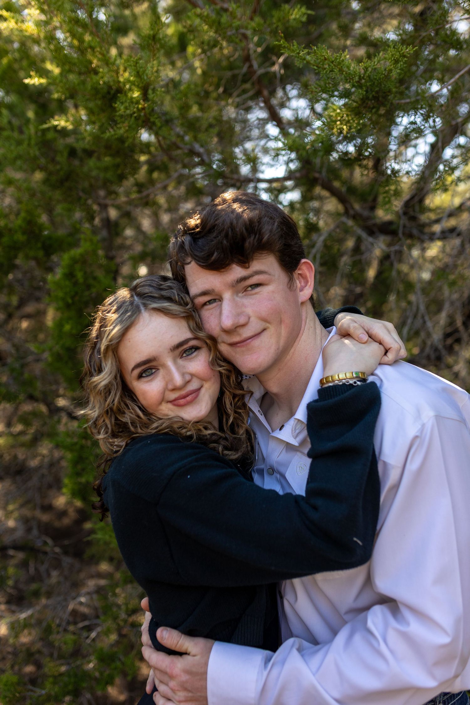 Two people sharing a tender embrace outdoors with trees and natural lighting in the background.