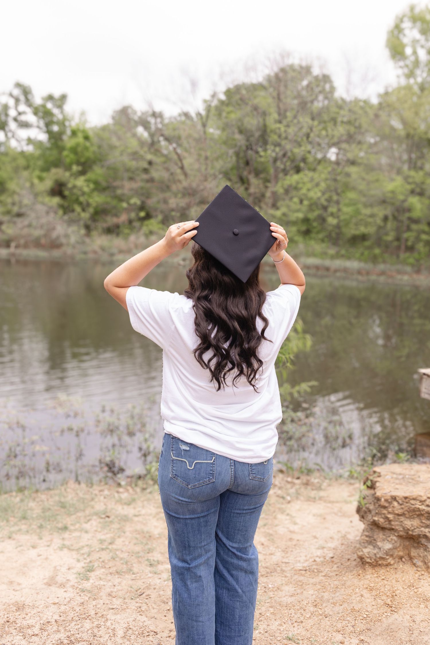 A person in casual attire and graduation cap stands near a lake surrounded by trees.