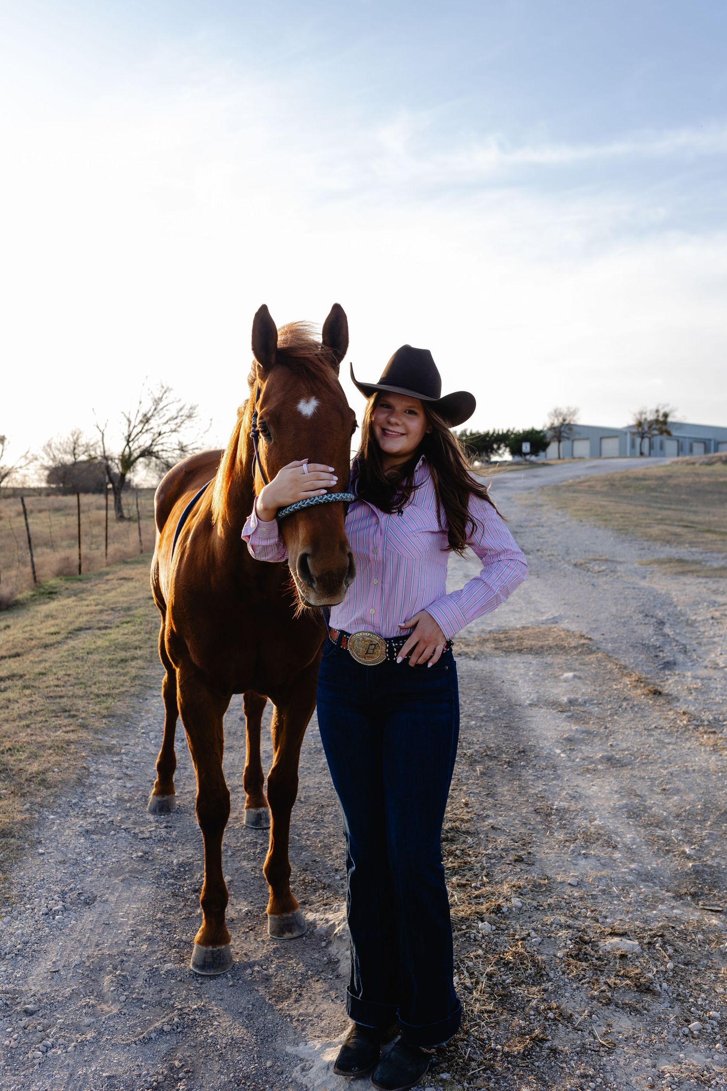 A brown horse stands with someone in western attire on a dirt path during sunset.