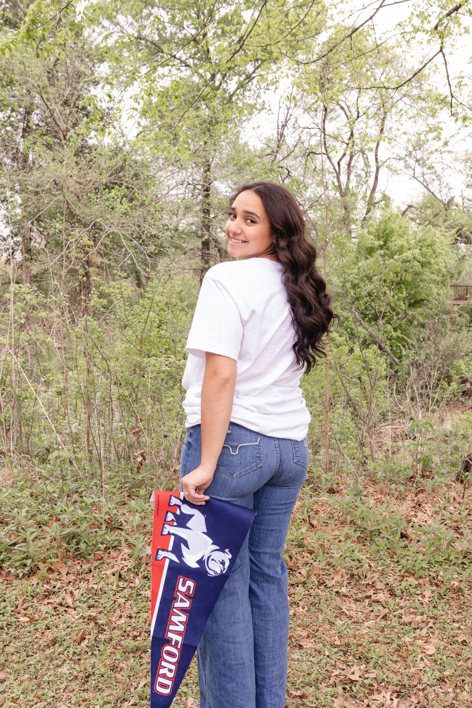 A person wearing a white t-shirt and jeans holds a blue Sam's Club shopping bag in a wooded area.