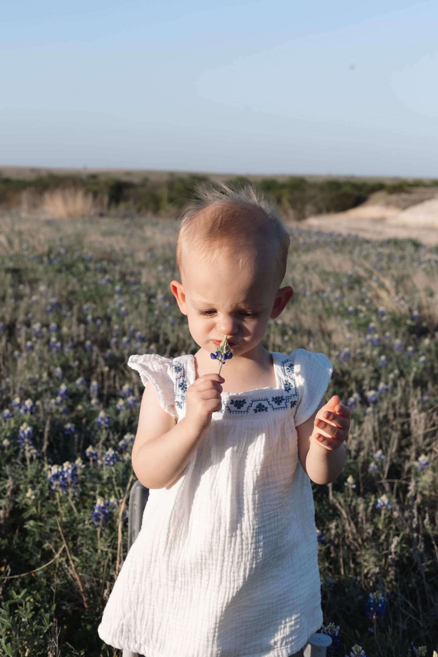 A small child in a white sleeveless dress stands among blooming wildflowers in a field at sunset.
