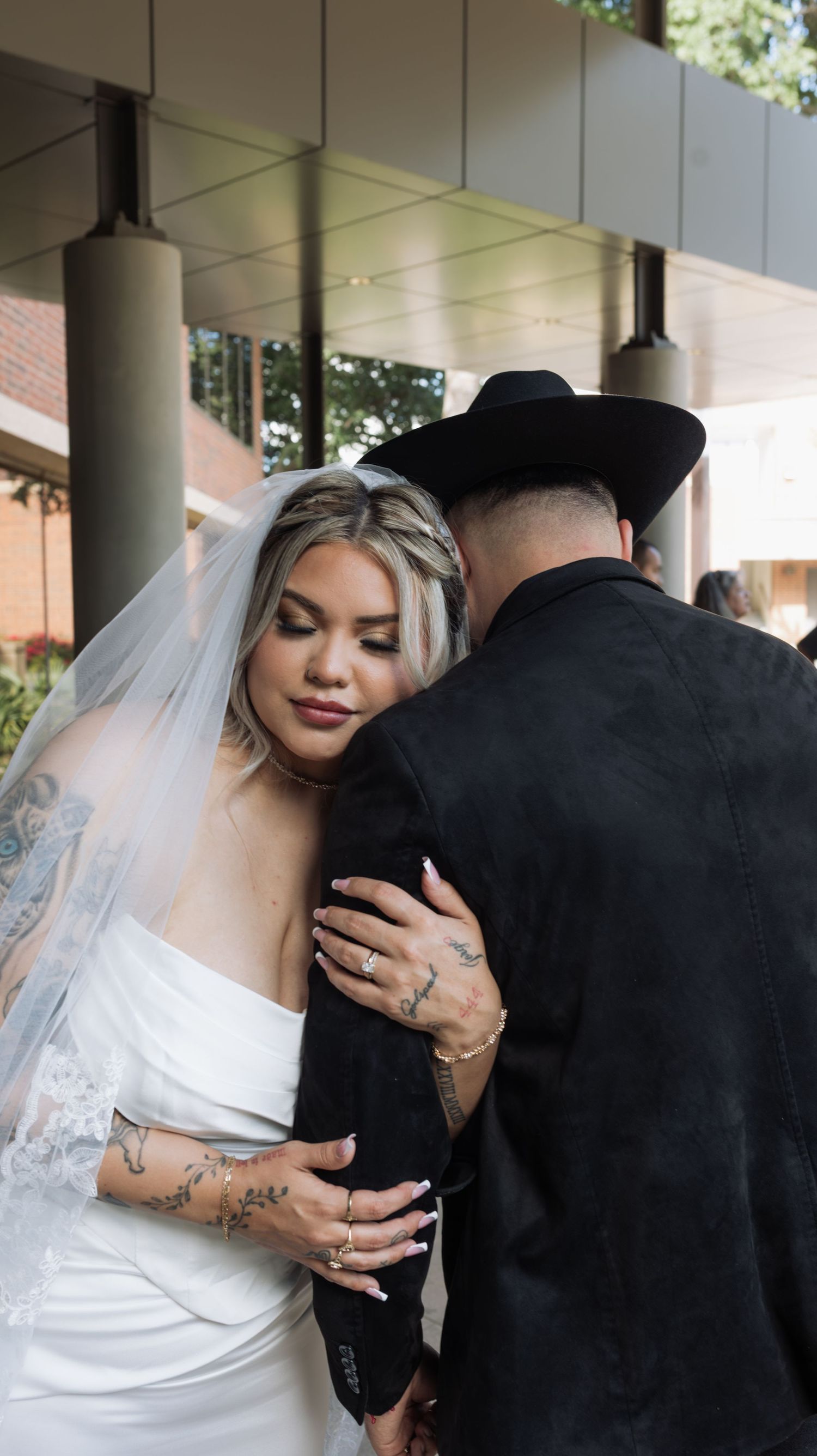 A bride in a white gown and veil embraces a groom in a black suit and cowboy hat at their wedding ceremony.