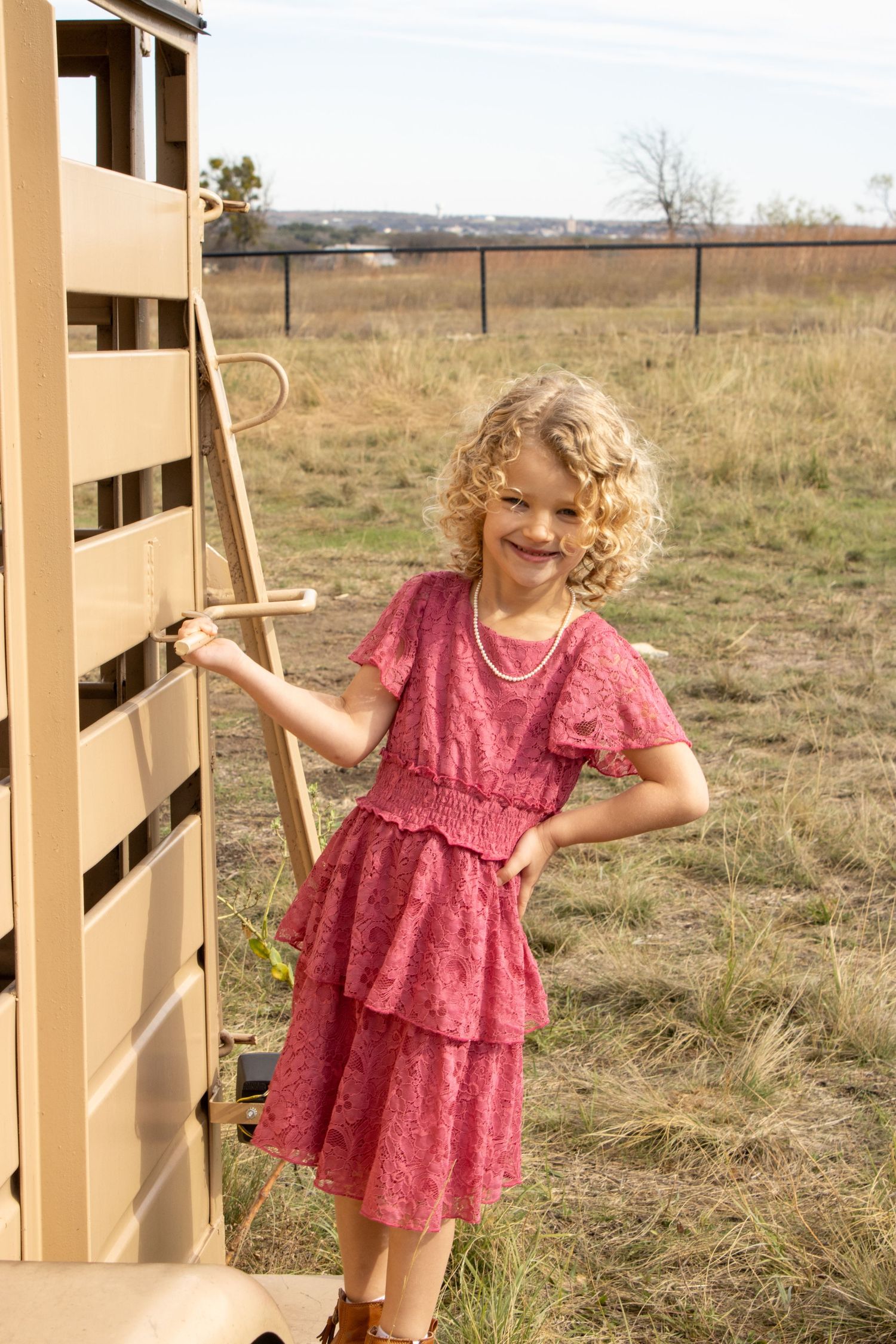 Young child in a pink lace dress plays near a wooden fence in a rural field setting during golden hour.