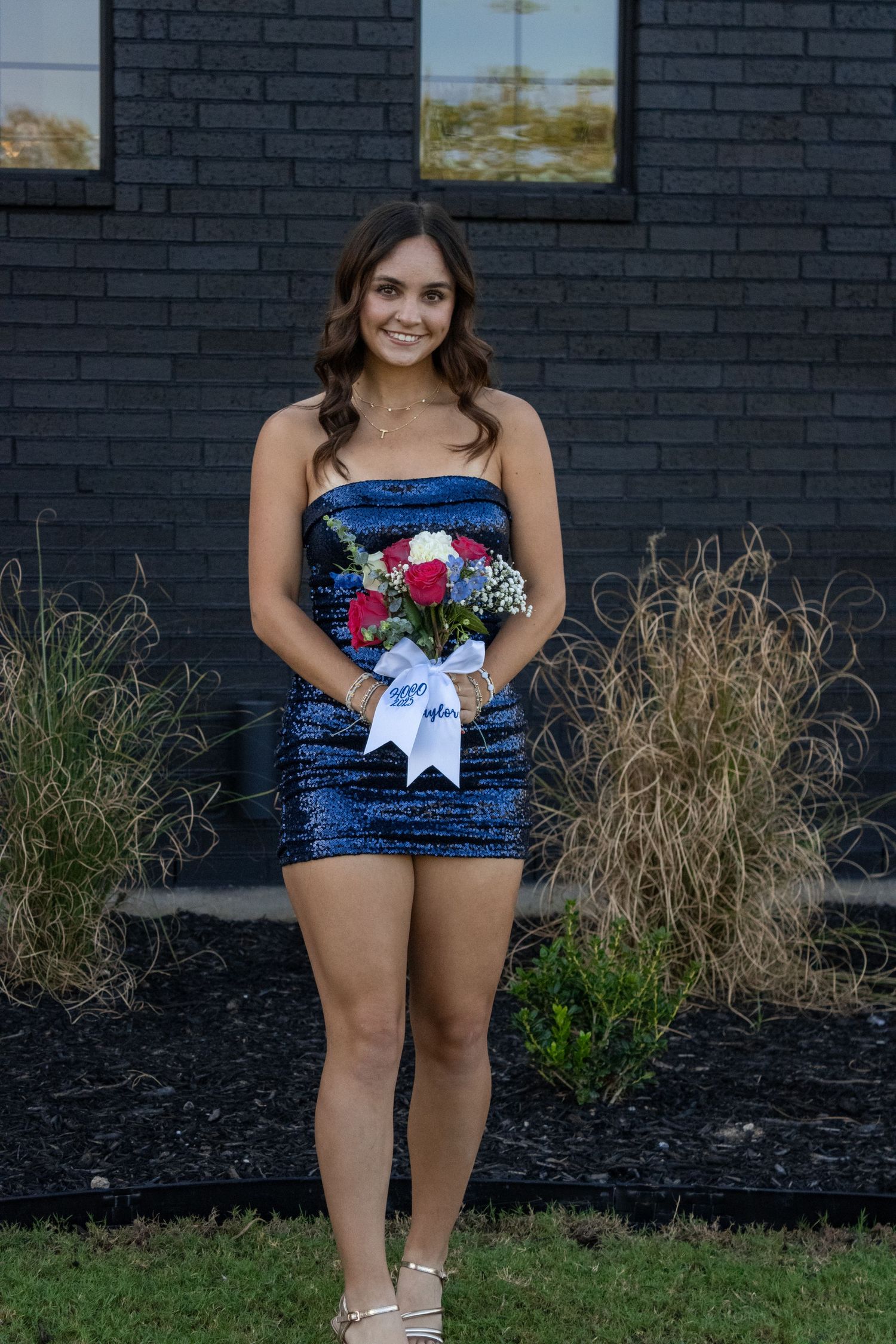 Portrait against navy brick wall wearing sparkly blue dress with red and white floral accents.