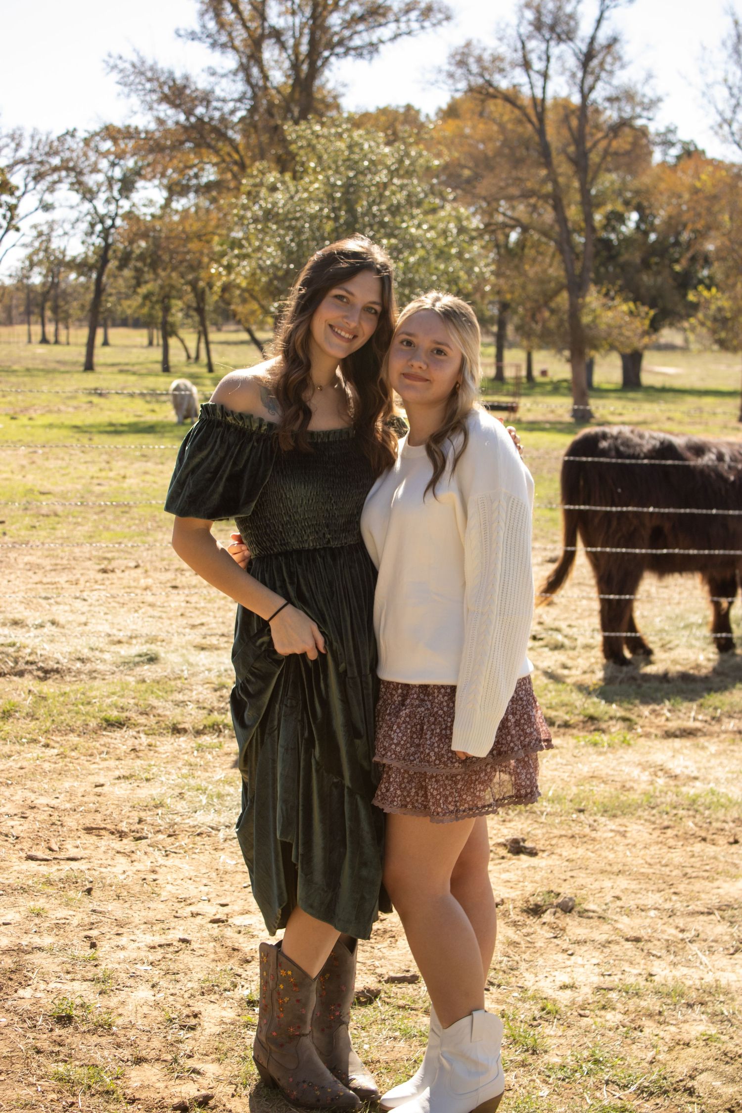 Two friends pose together in a sunny autumn field with bison grazing in the background.