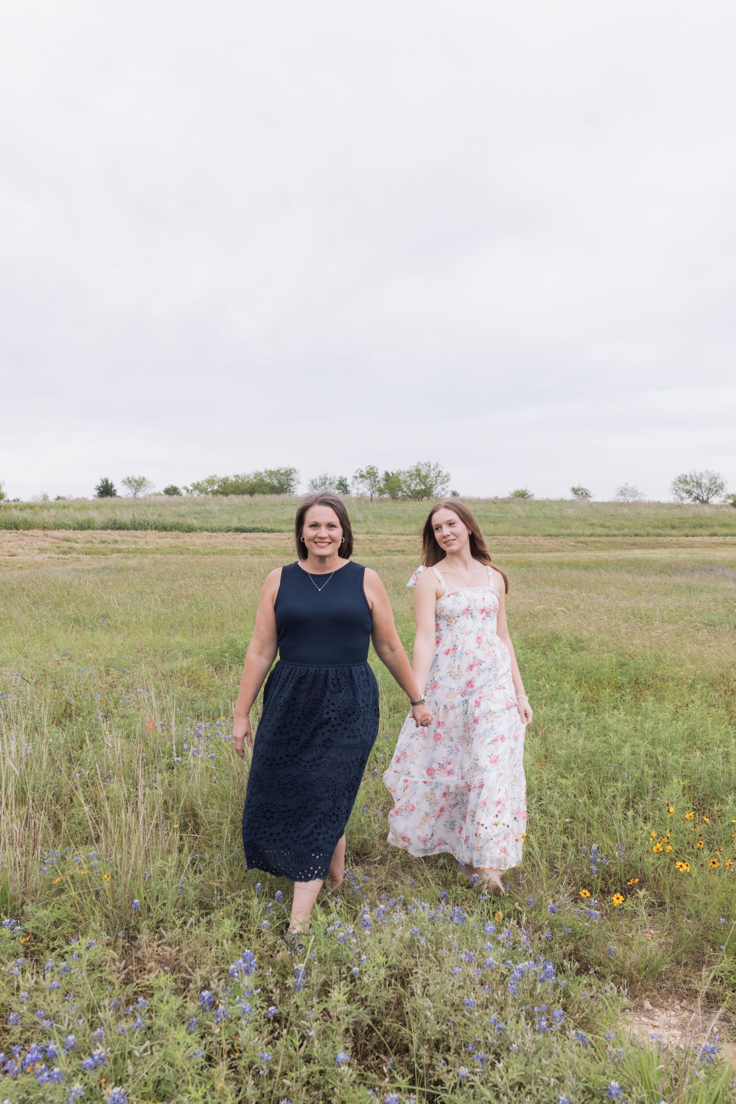 Two women in elegant dresses hold hands while walking through a blooming wildflower field on an overcast day.