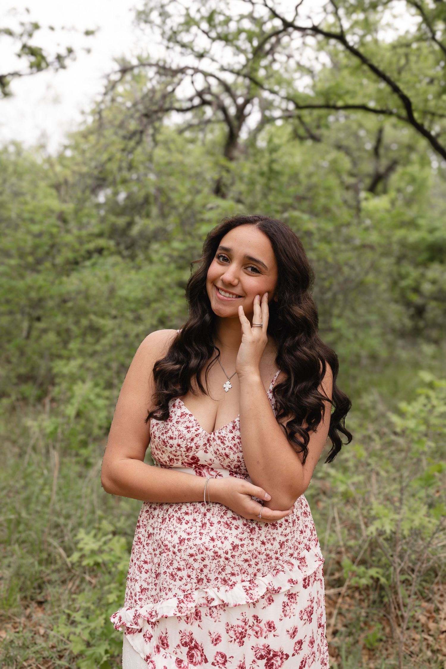 A person in a red and white floral dress poses playfully in a natural outdoor setting with trees and greenery.