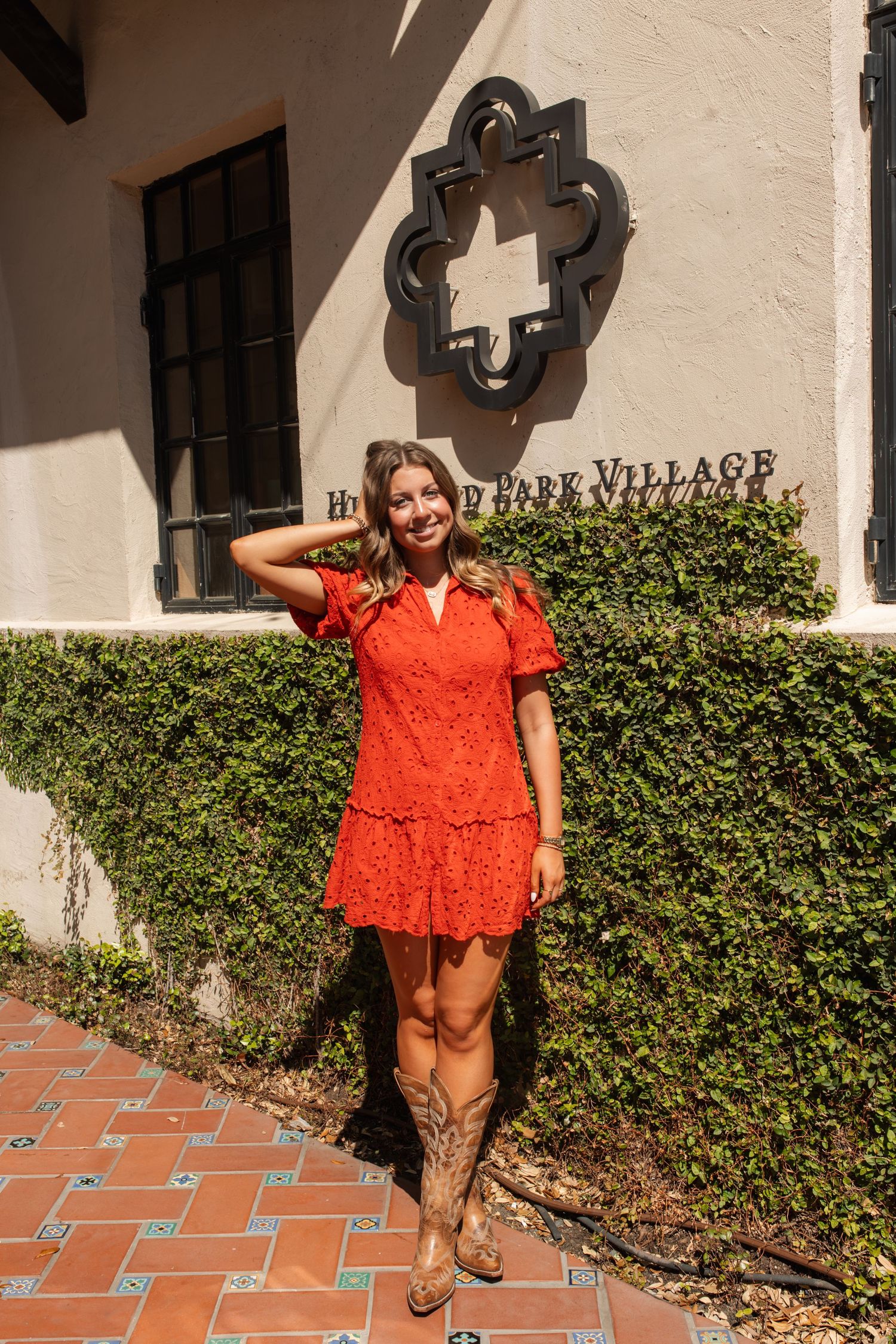 A vibrant coral-colored eyelet lace dress styled with brown sandals against a Spanish-style building exterior.