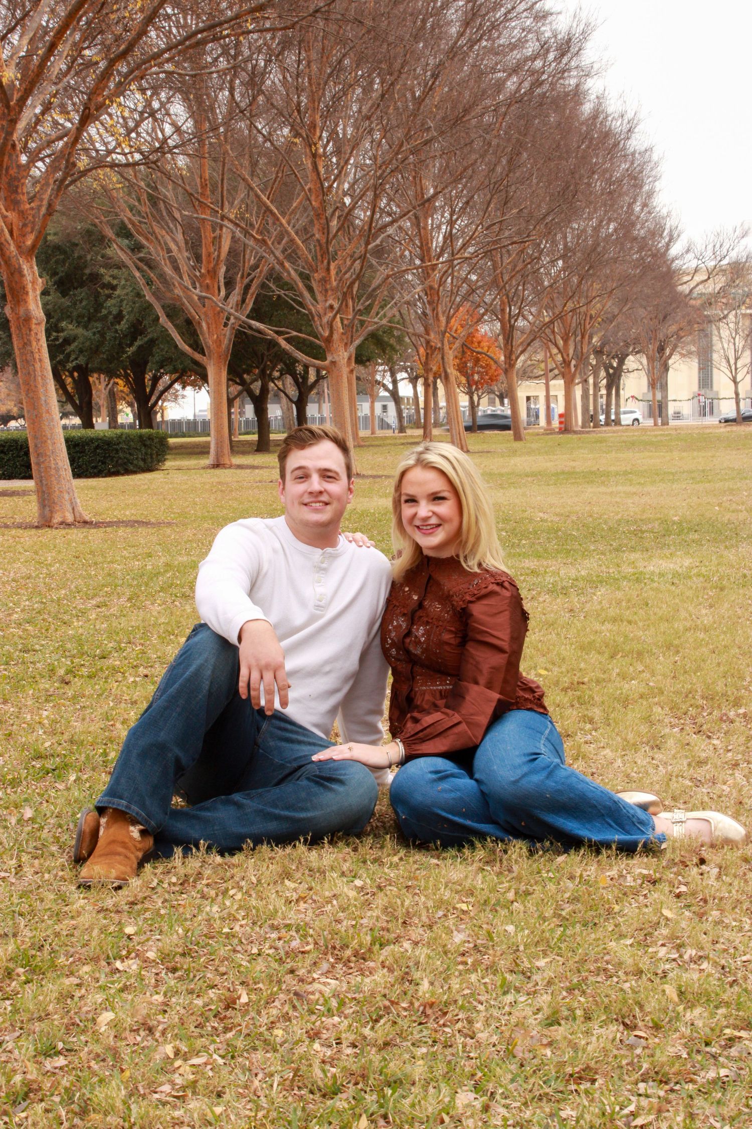 A couple relaxes on grass beneath a row of bare winter trees on a sunny day.