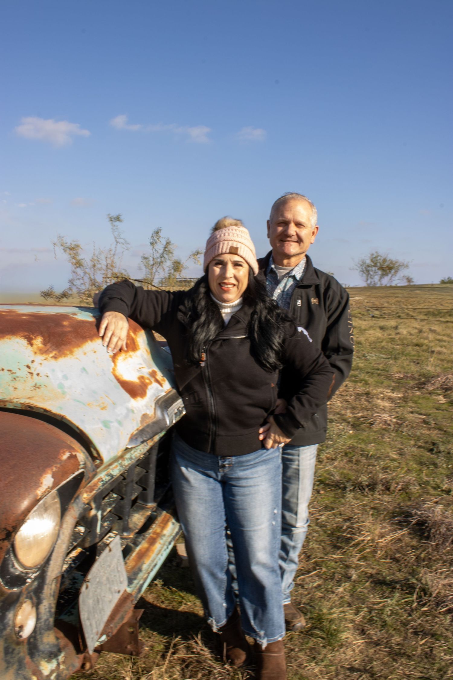 A couple poses together next to a rusty vintage car in an open field on a sunny day.
