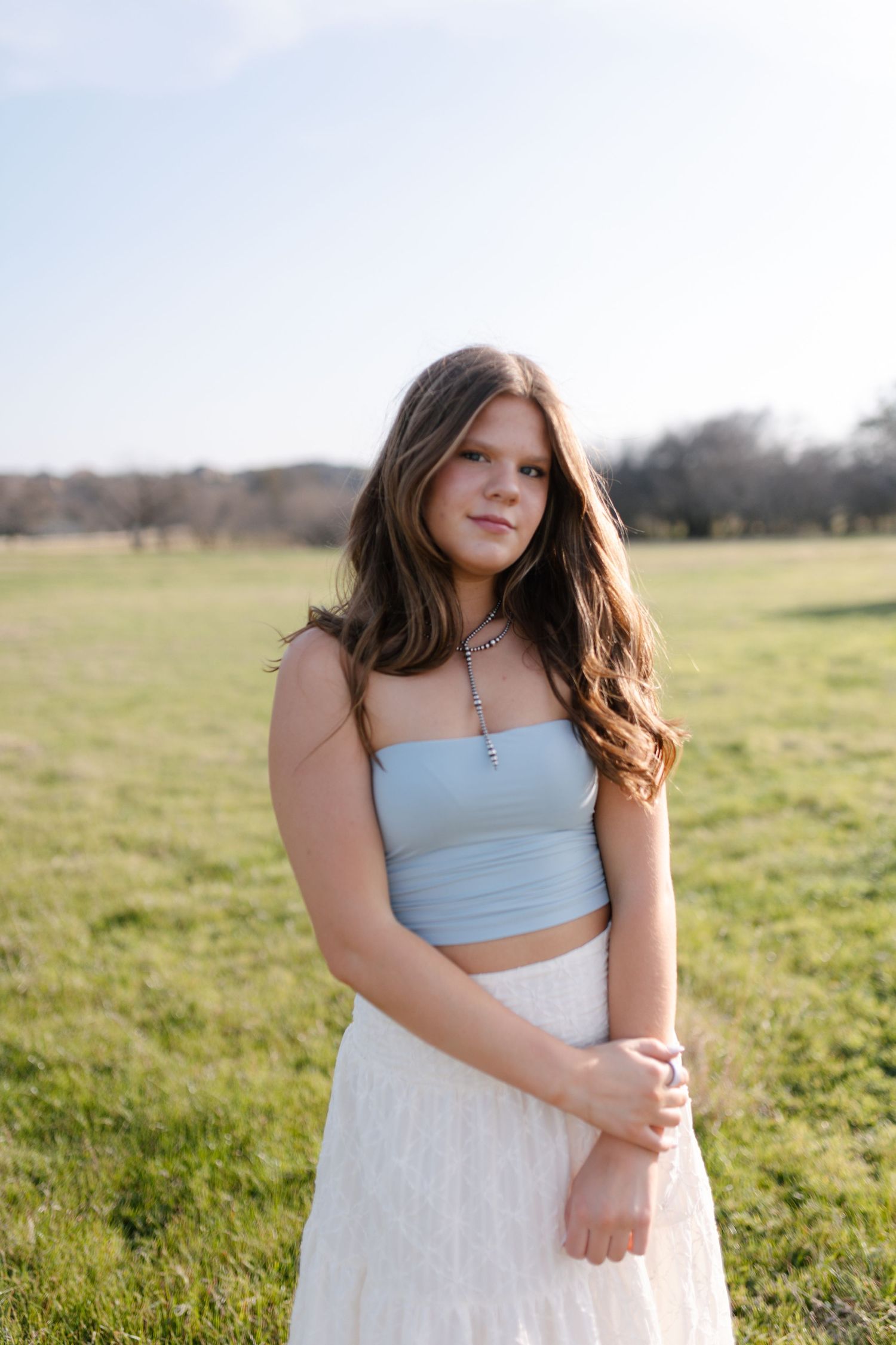 A soft portrait in a sunlit field wearing a light blue top and white lace skirt against a natural outdoor backdrop.