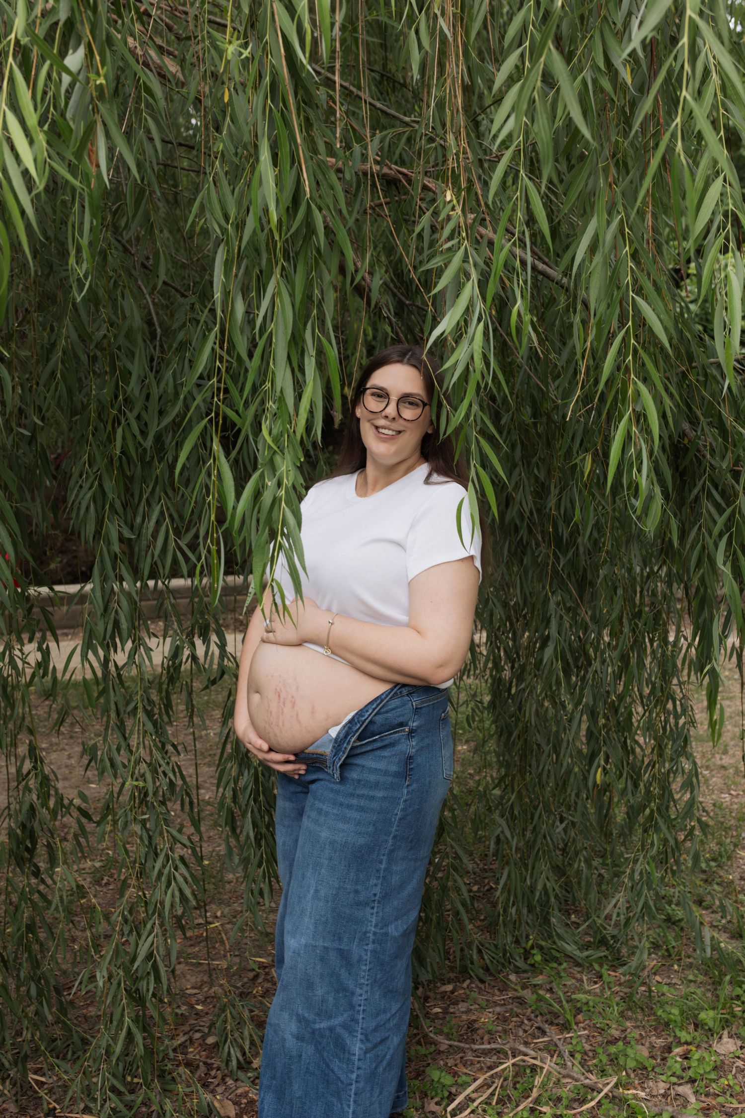 Smiling pregnant woman in white crop top and jeans cradling her baby bump outdoors under weeping willow tree.