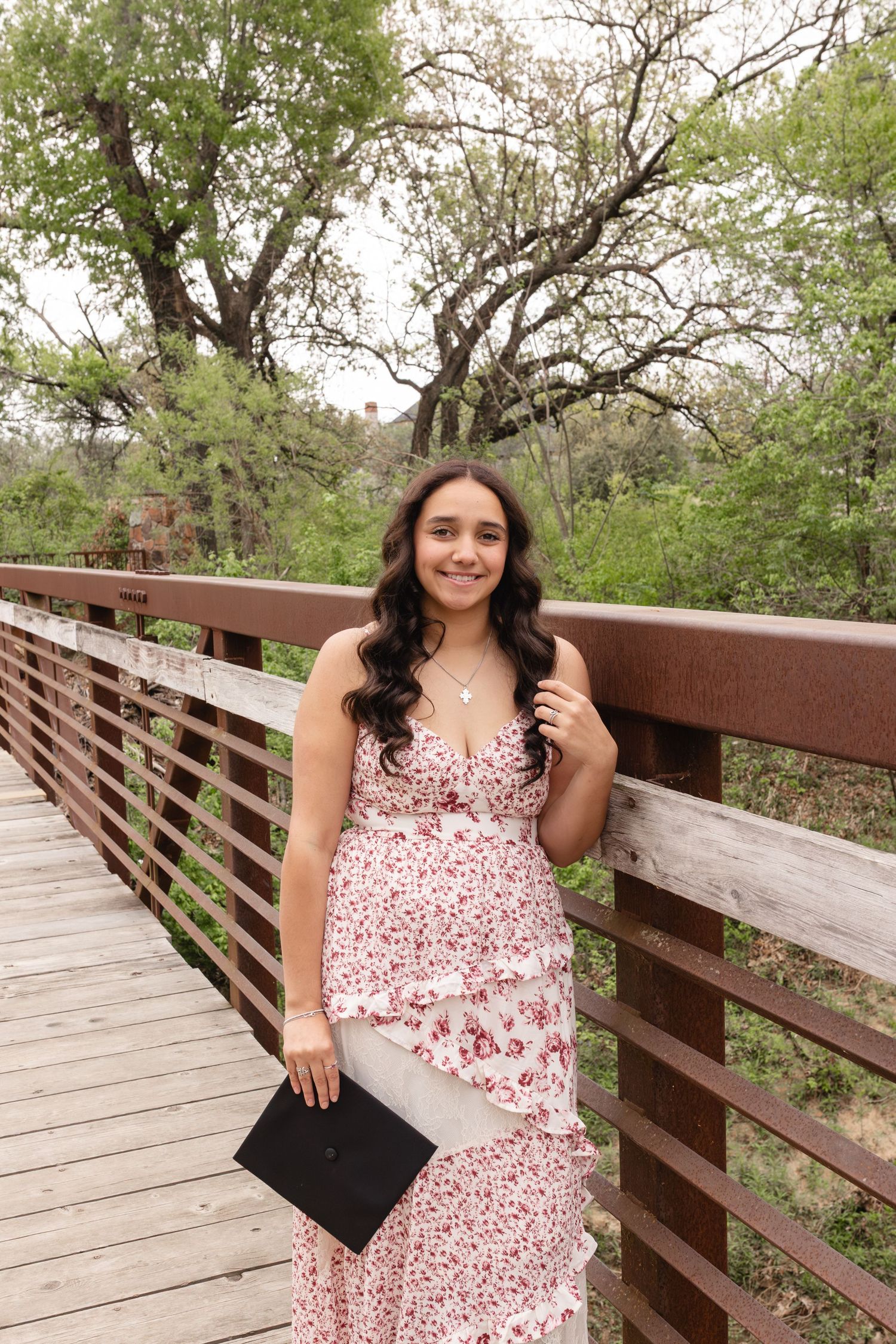 Young woman in floral dress holding graduation cap on wooden bridge surrounded by lush green trees.