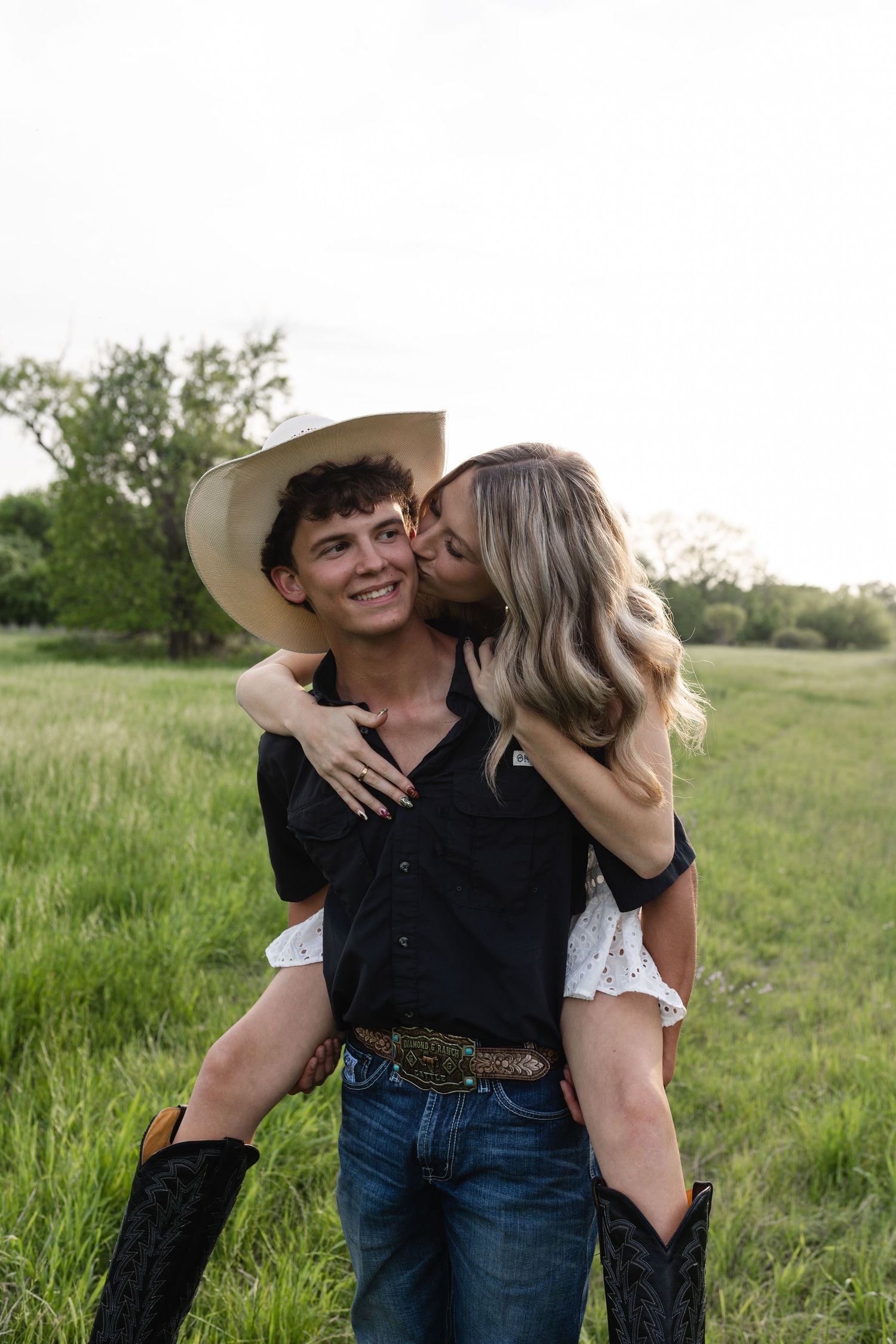 A woman kisses a smiling cowboy giving her a piggyback ride in a lush green field.
