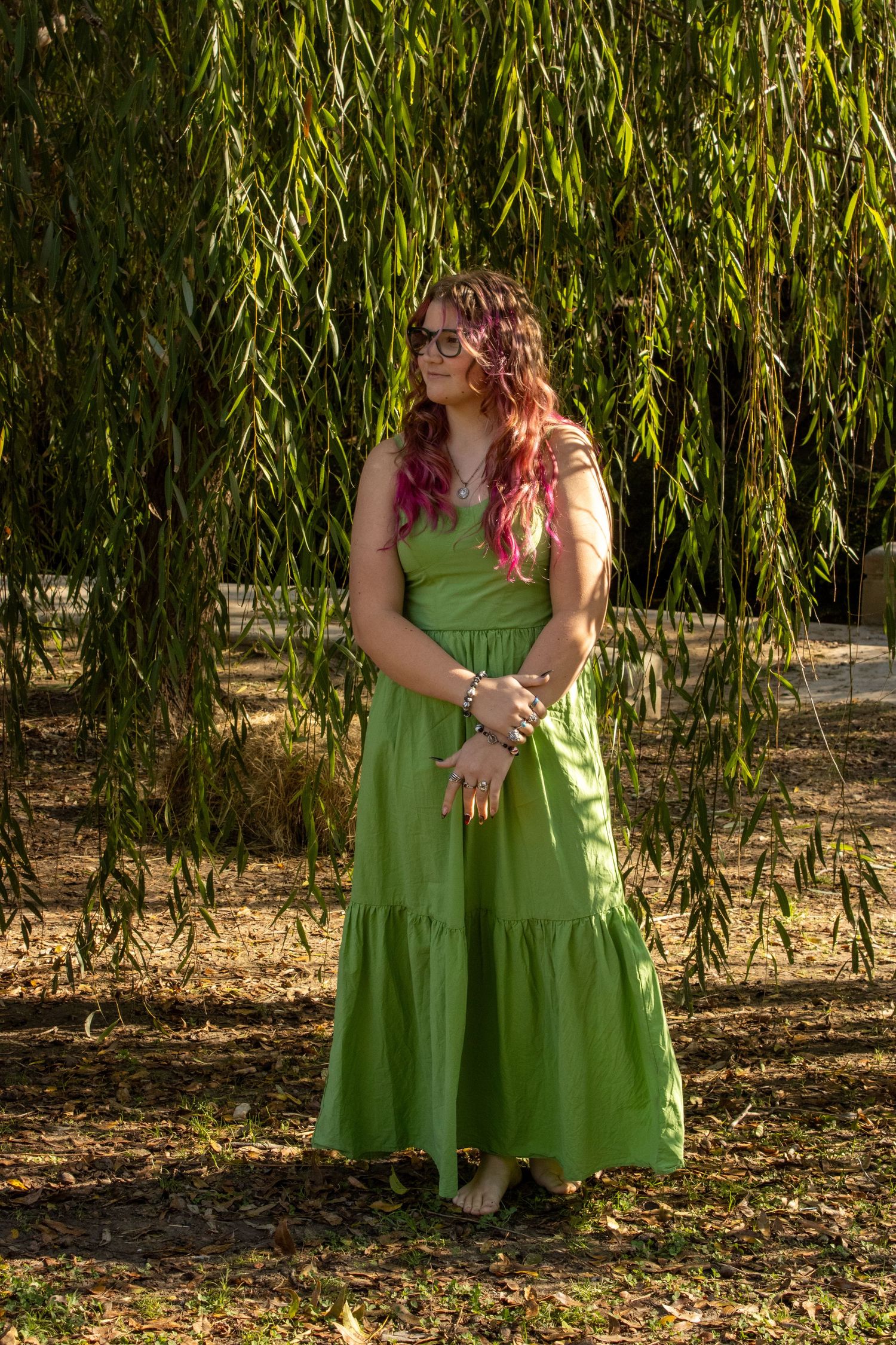 Two people in matching green dresses sit beneath a weeping willow tree in a park setting.