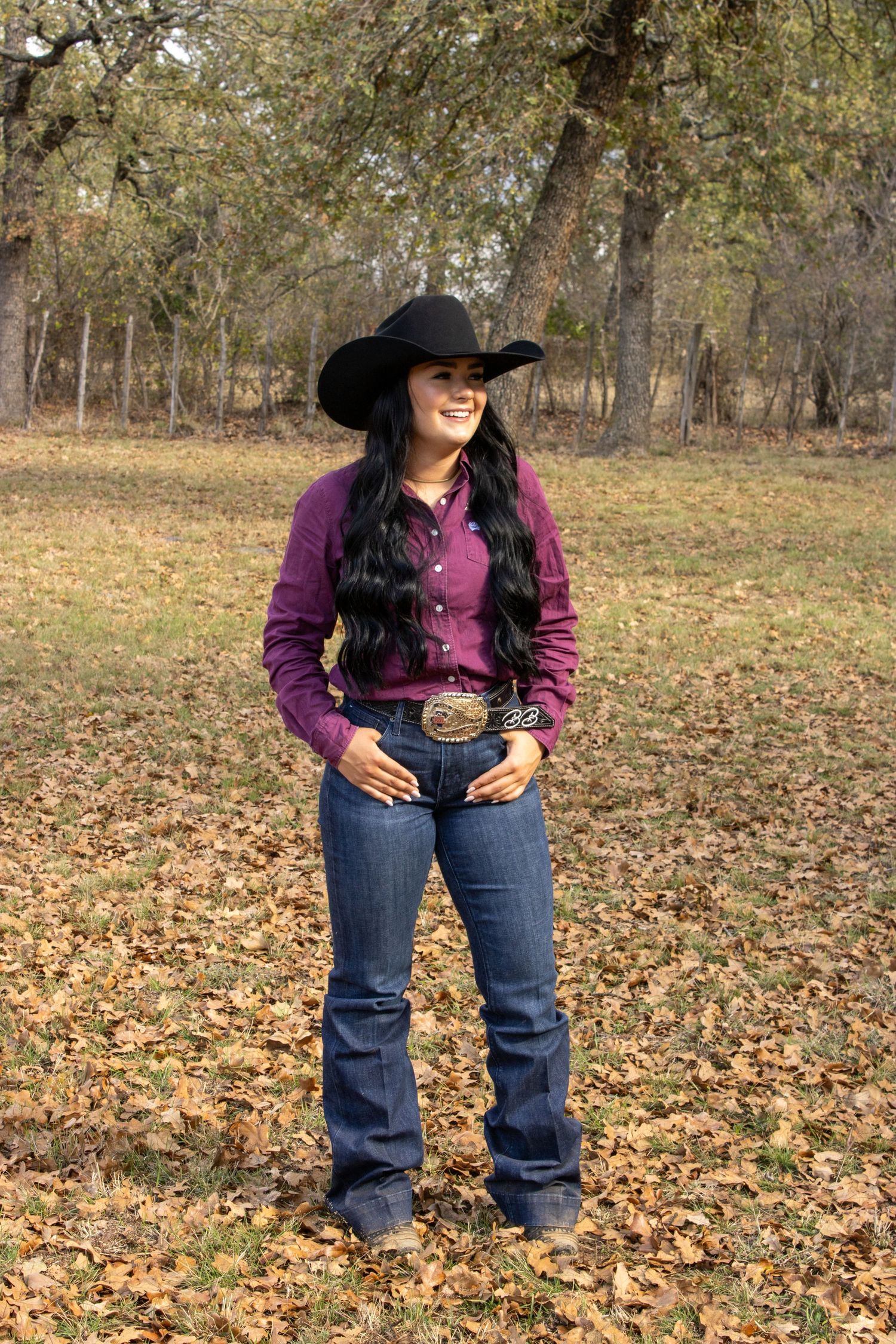 A person wearing western attire and a black hat stands in a field of autumn leaves.
