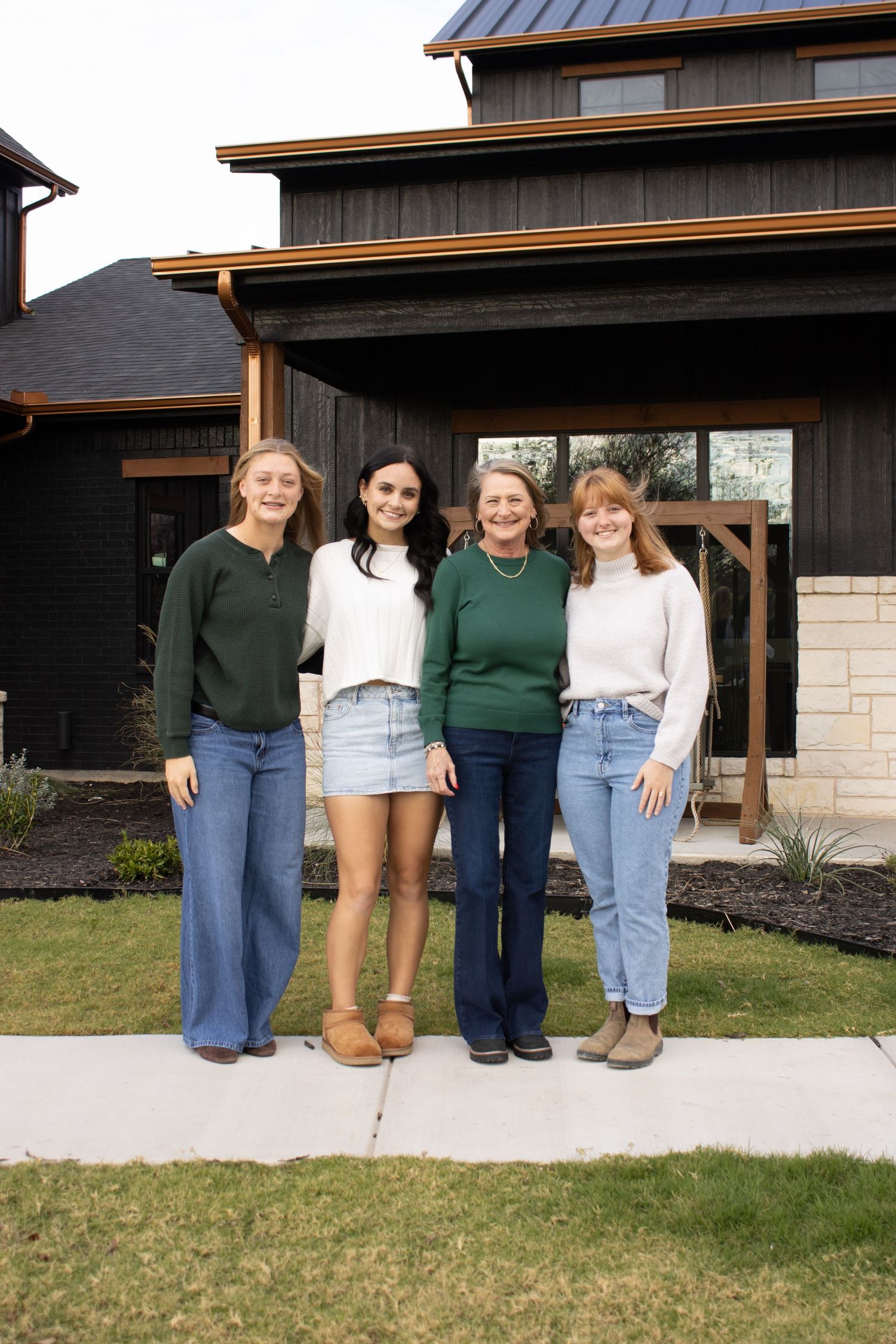 Four people wearing casual outfits stand together in front of a modern house with dark siding and stone accents.