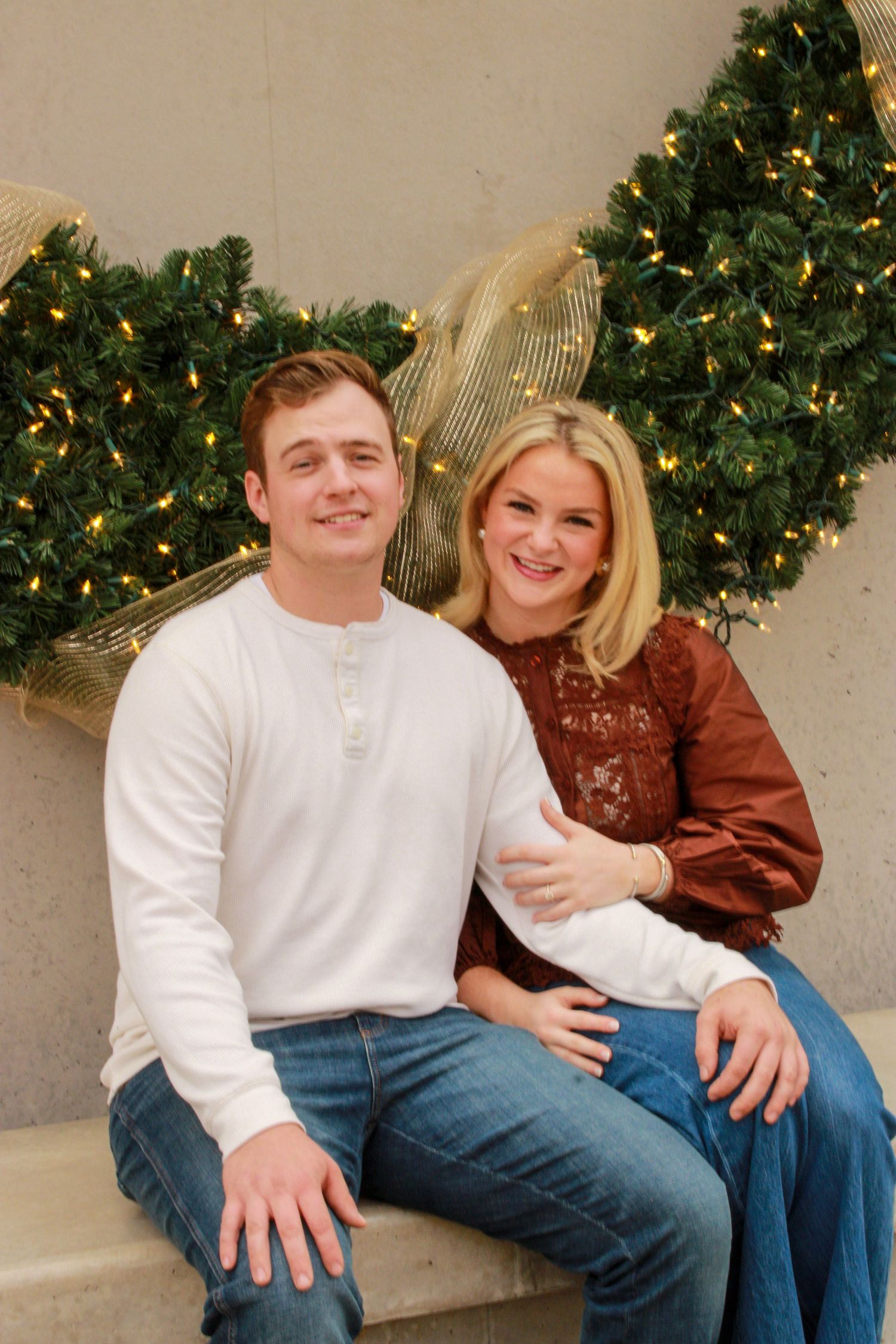 A couple sits together on a stone bench in front of a decorated Christmas tree wearing casual winter clothing.