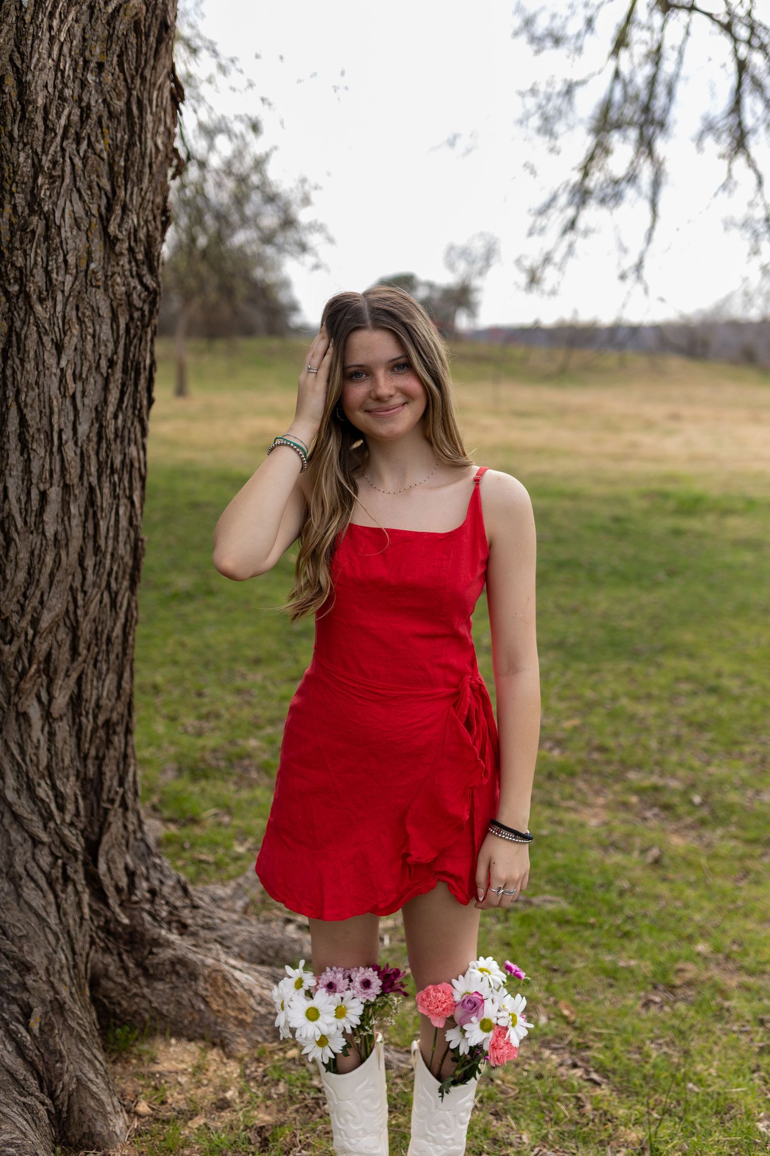 A person in a red dress poses outdoors near a tree trunk with white and pink flowers in a rustic setting.