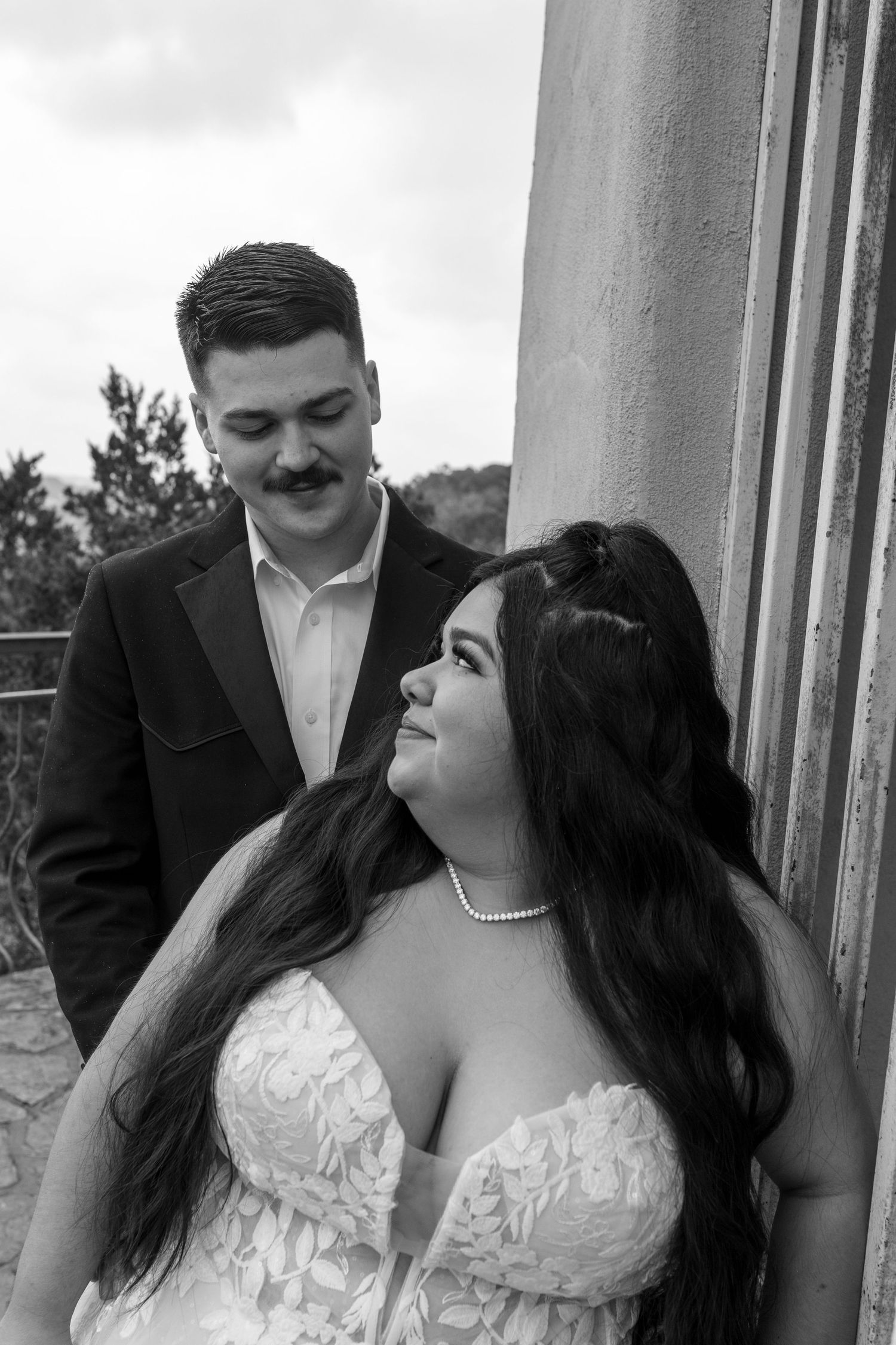 Black and white wedding photo of a bride in lace gown with pearl necklace leaning against column as groom in suit looks down at her.