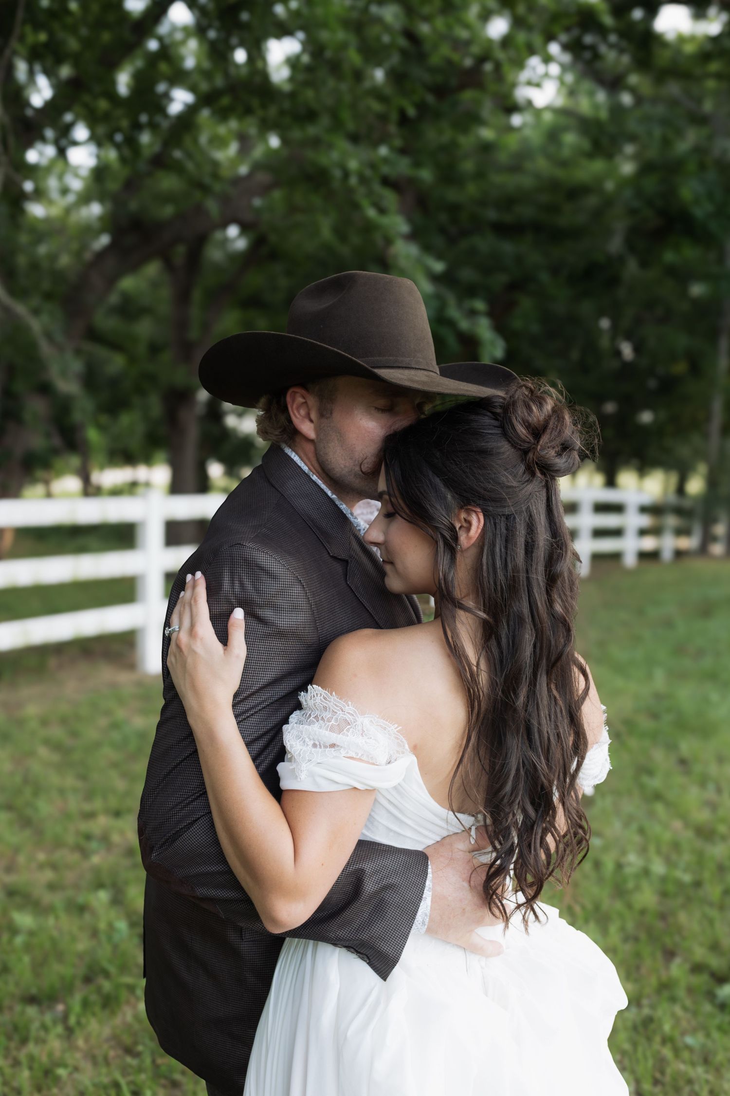 Groom in cowboy hat embraces bride in off-shoulder white gown near white fence in outdoor setting.