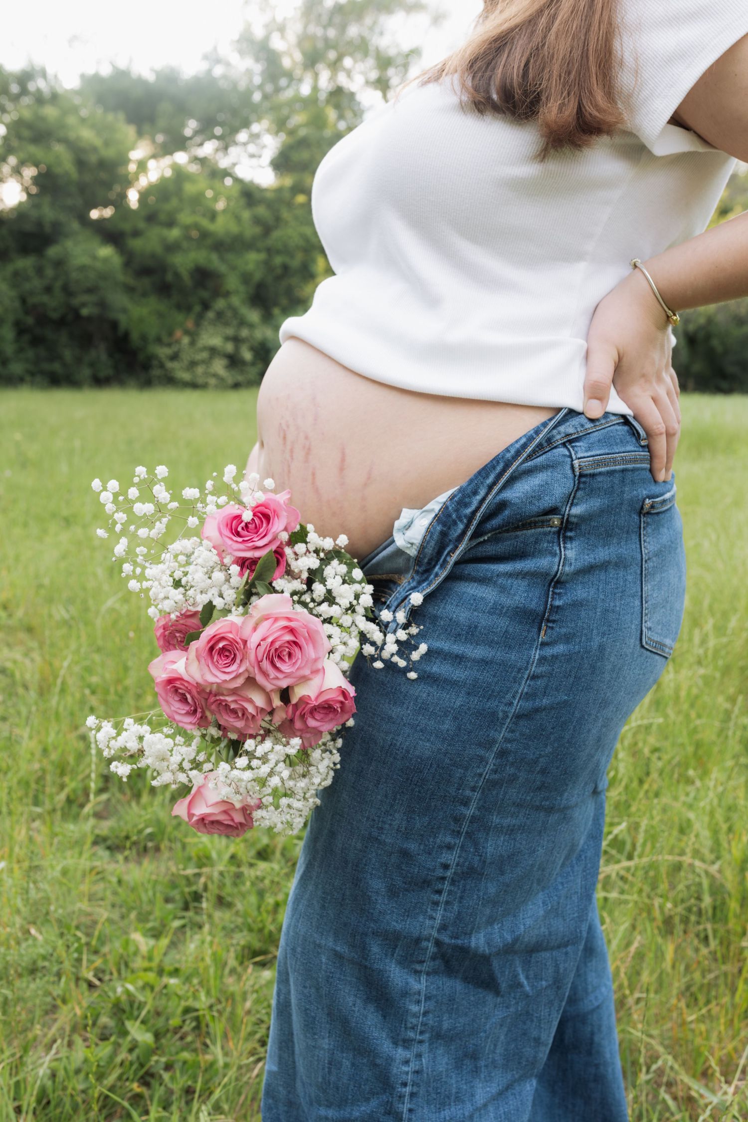 Pregnant woman outdoors showing bare belly with pink roses and baby's breath tucked into unbuttoned jeans.