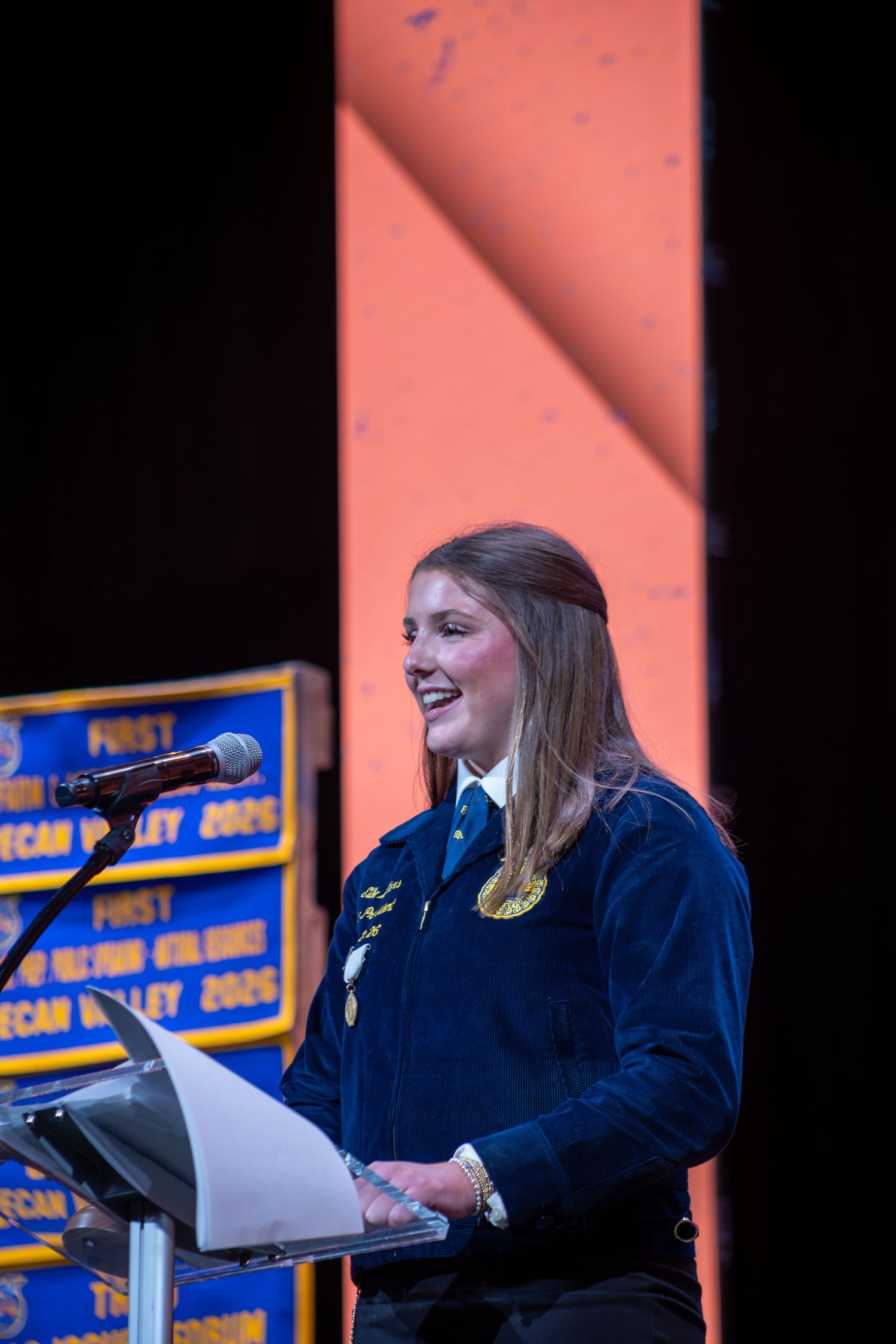 Young woman in FFA blue jacket speaks at podium with gold medal, smiling on stage at FFA event.