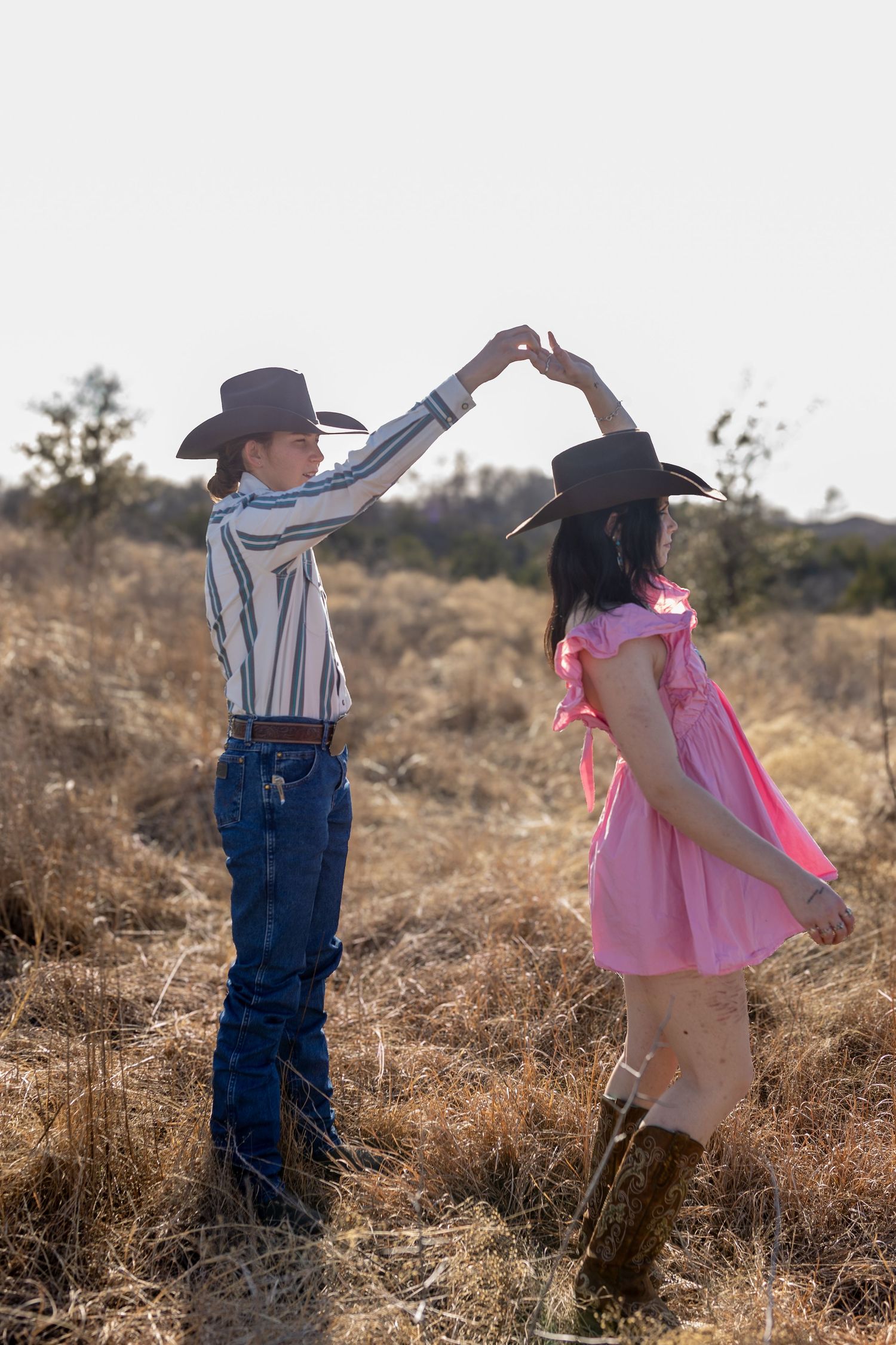 Two young children in cowboy attire dance together in a sunlit field during golden hour.