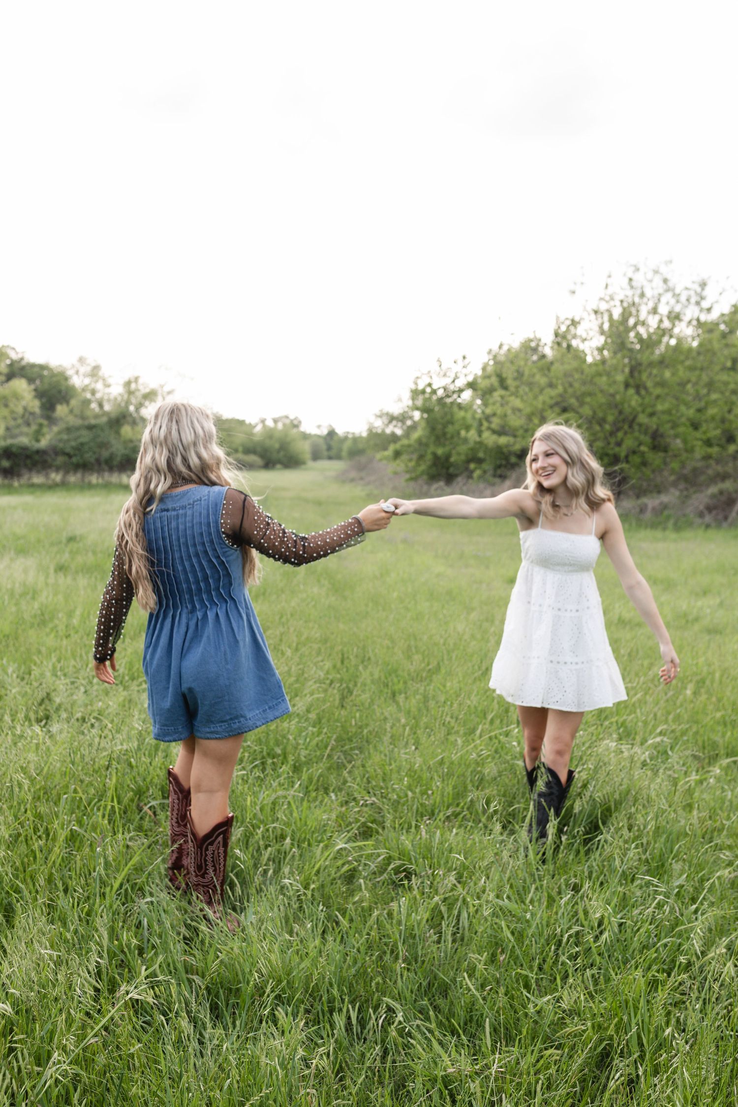 Two blonde women in cowboy boots hold hands dancing in a green field, one in blue romper, one in white dress.