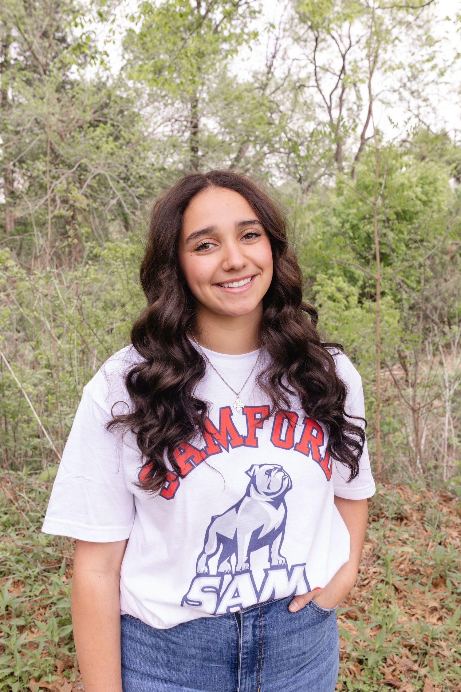 Young woman smiling outdoors in a Samford University white t-shirt, with long dark wavy hair, in a wooded setting.
