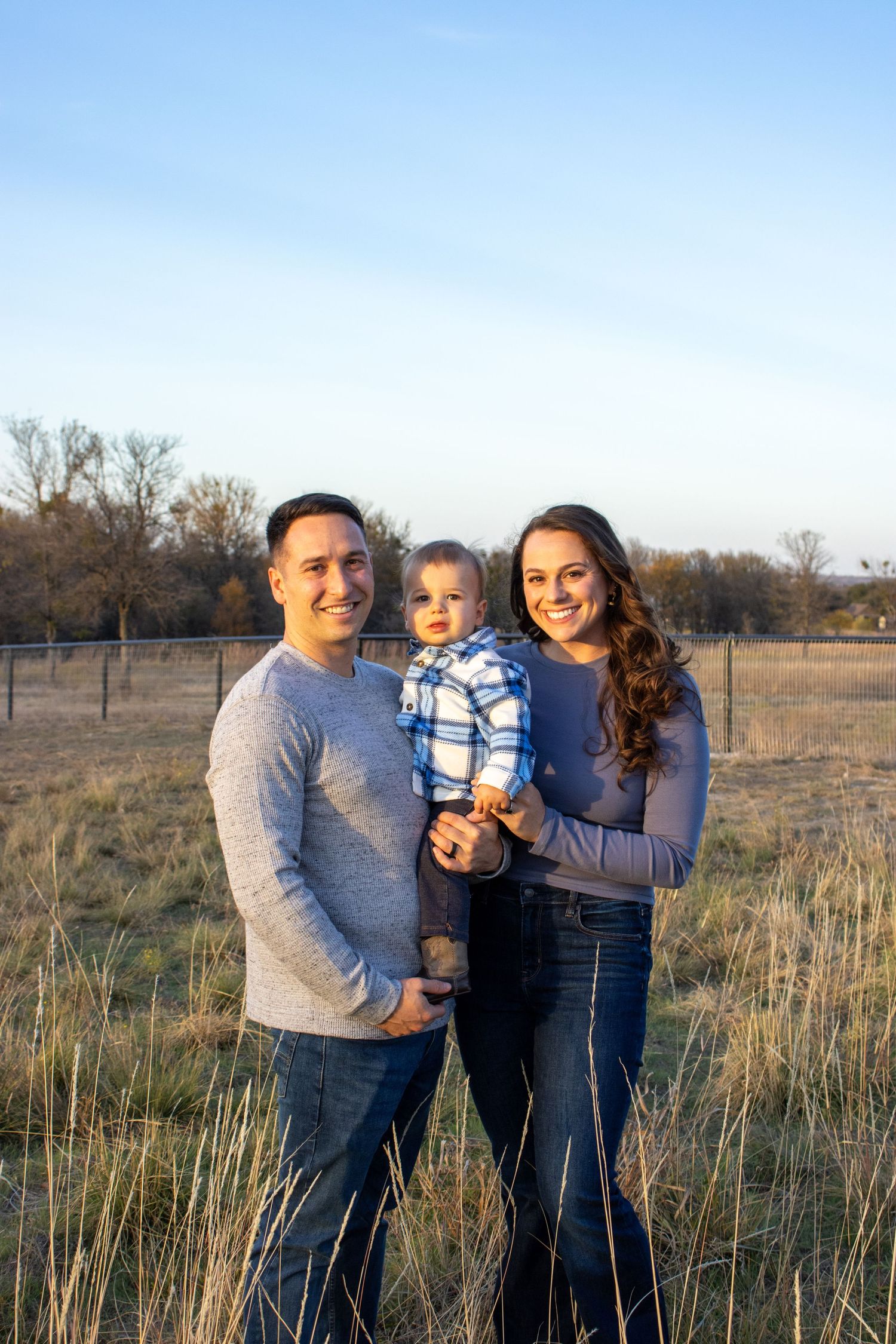 A family poses together in a rustic field setting during golden hour with tall grass and trees in the background.