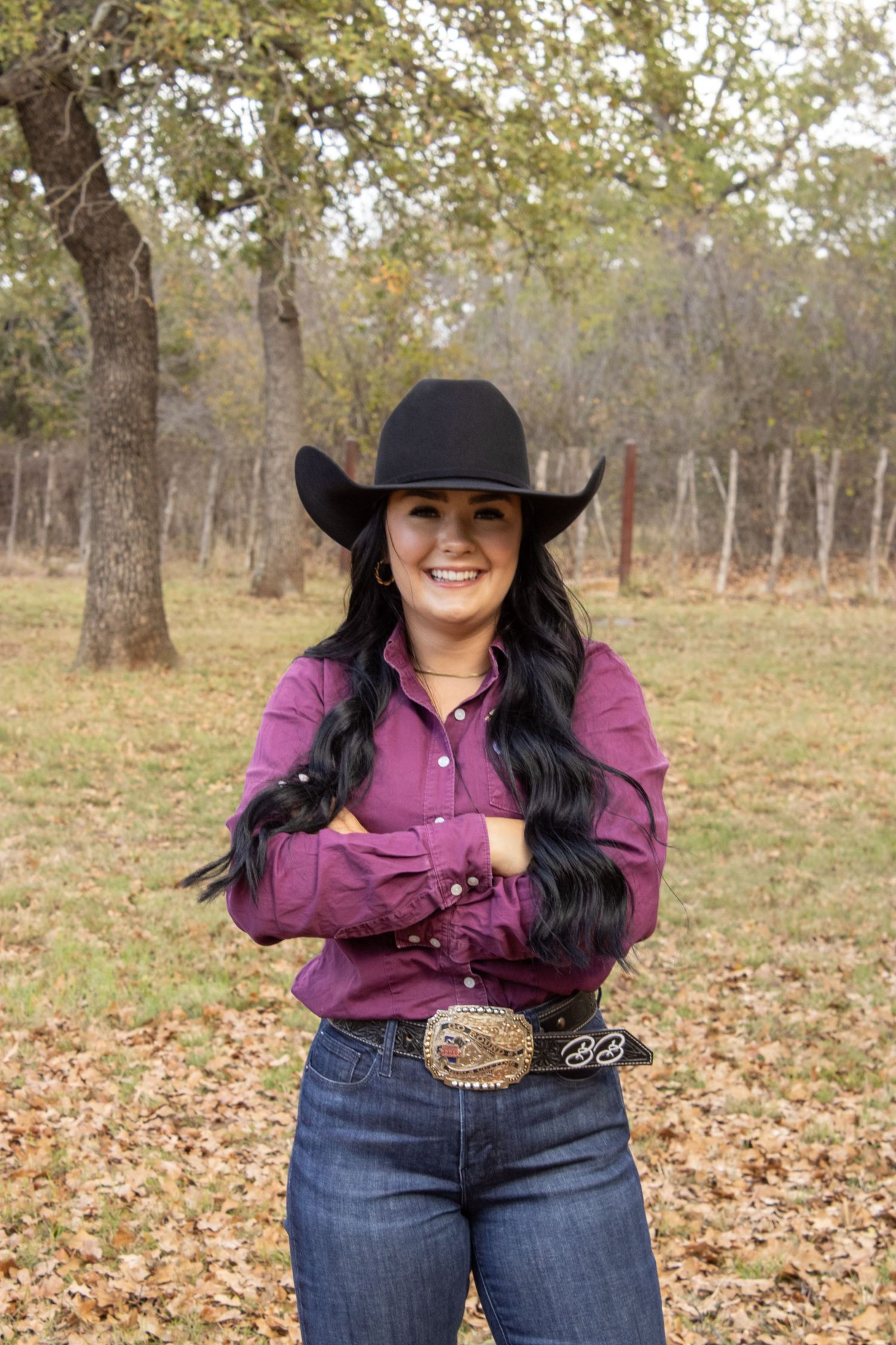 A person in western wear with a cowboy hat and purple button-down shirt poses outdoors among autumn trees.