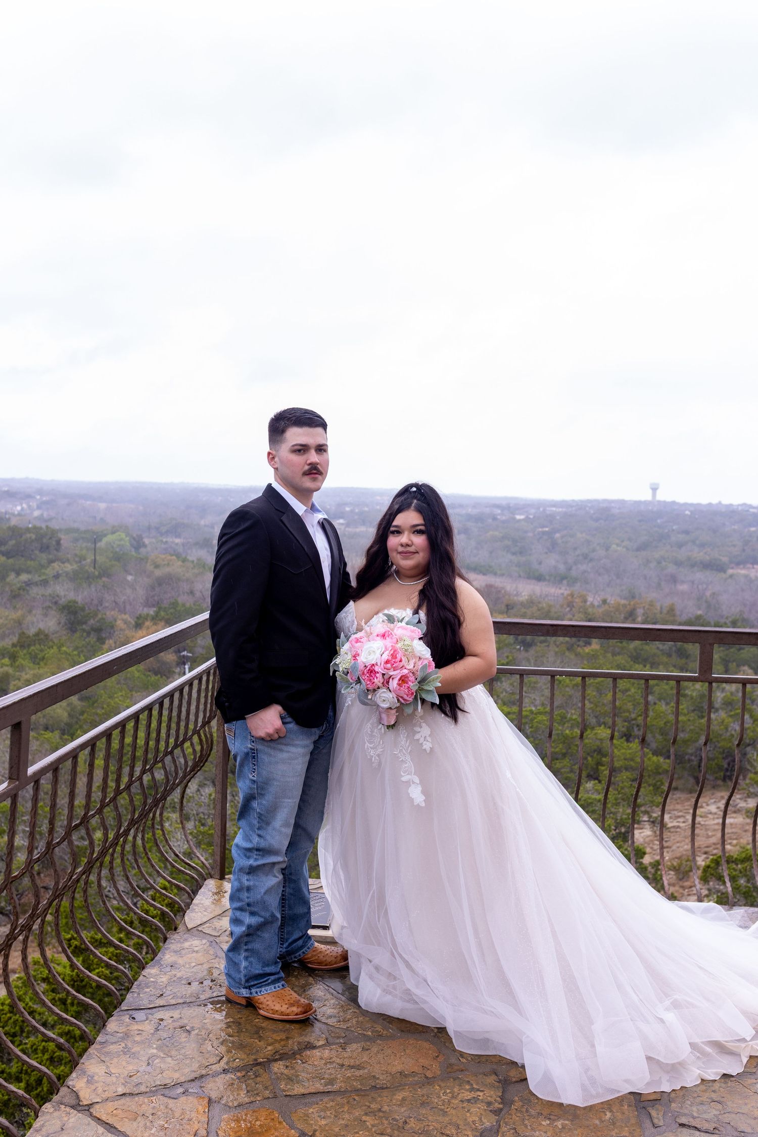 Bride in white ballgown holding pink bouquet poses with groom in black suit on scenic overlook balcony.