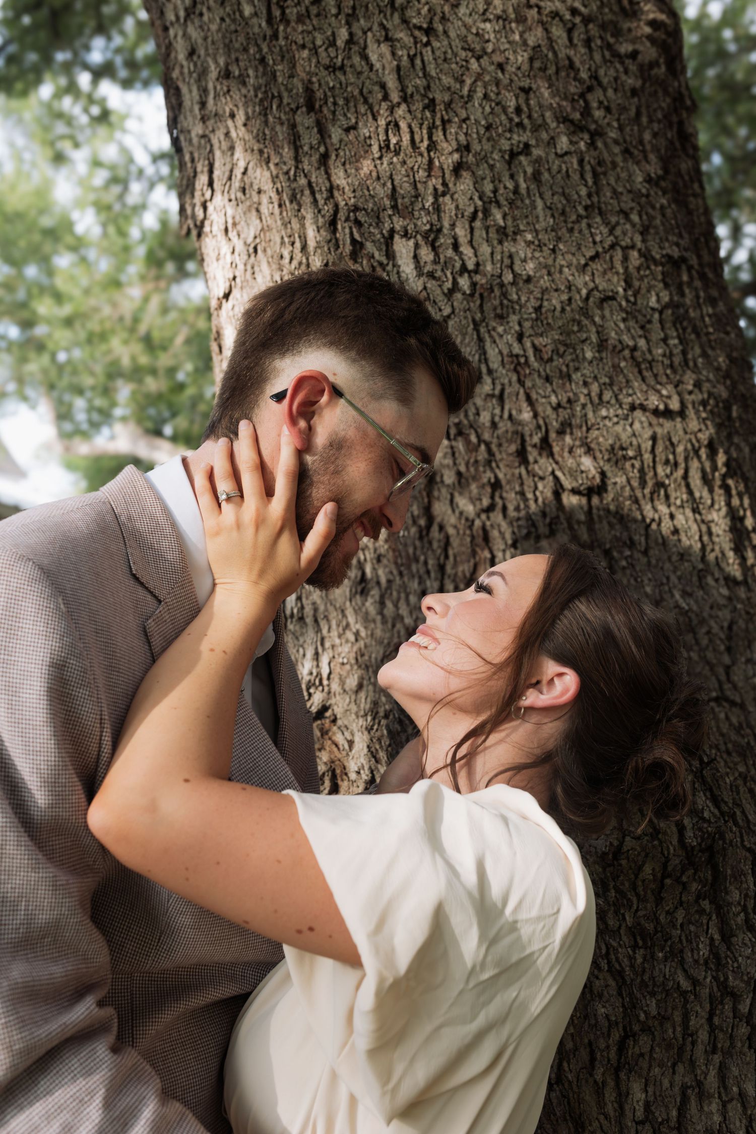 Romantic couple nearly kissing against a tree trunk outdoors, woman in white dress cradling mans face.