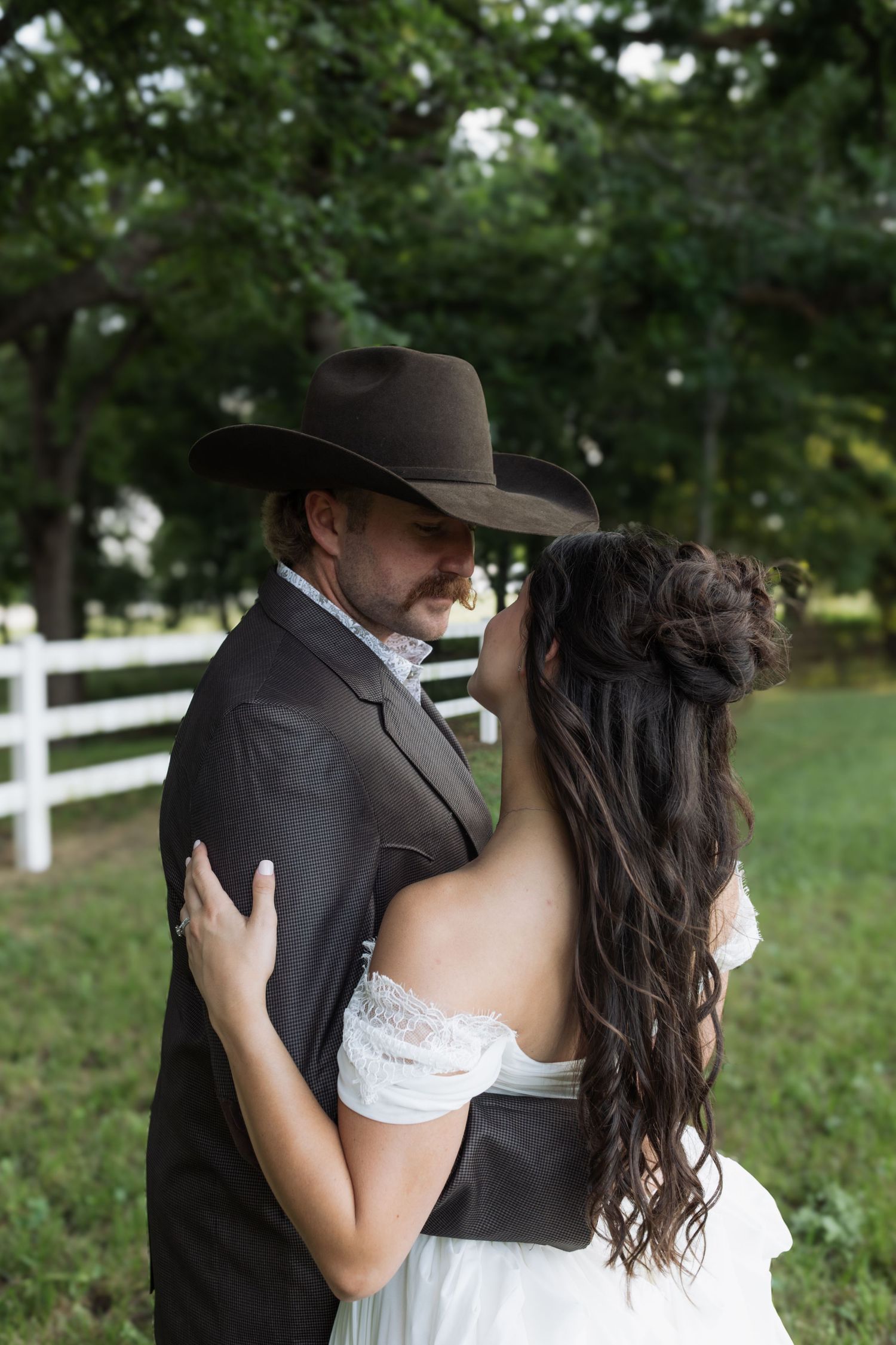 Bride and groom embracing near white fence at rustic ranch wedding in lush green setting.