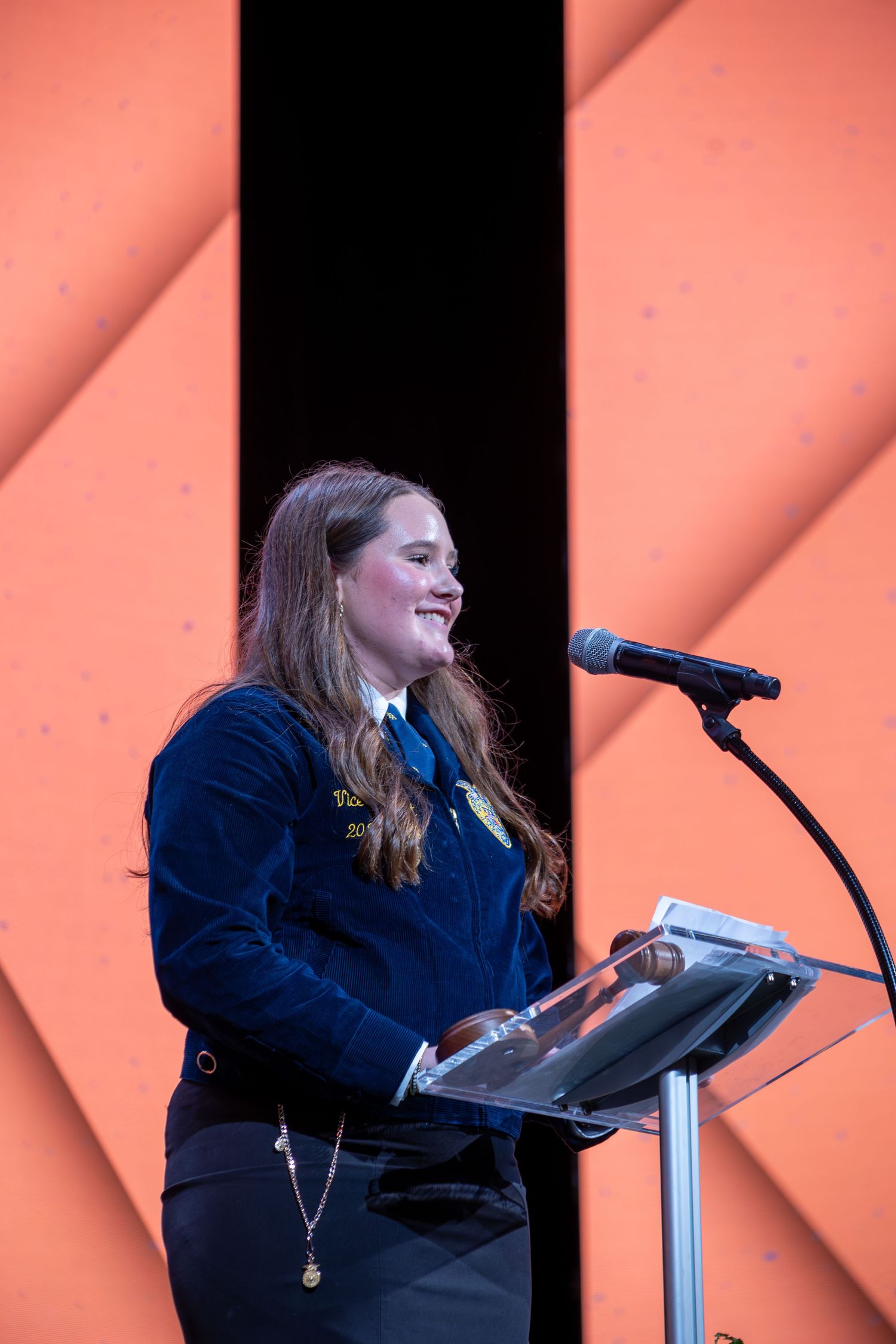 Young woman in FFA blue jacket speaks at a podium with microphone on stage with orange geometric backdrop.