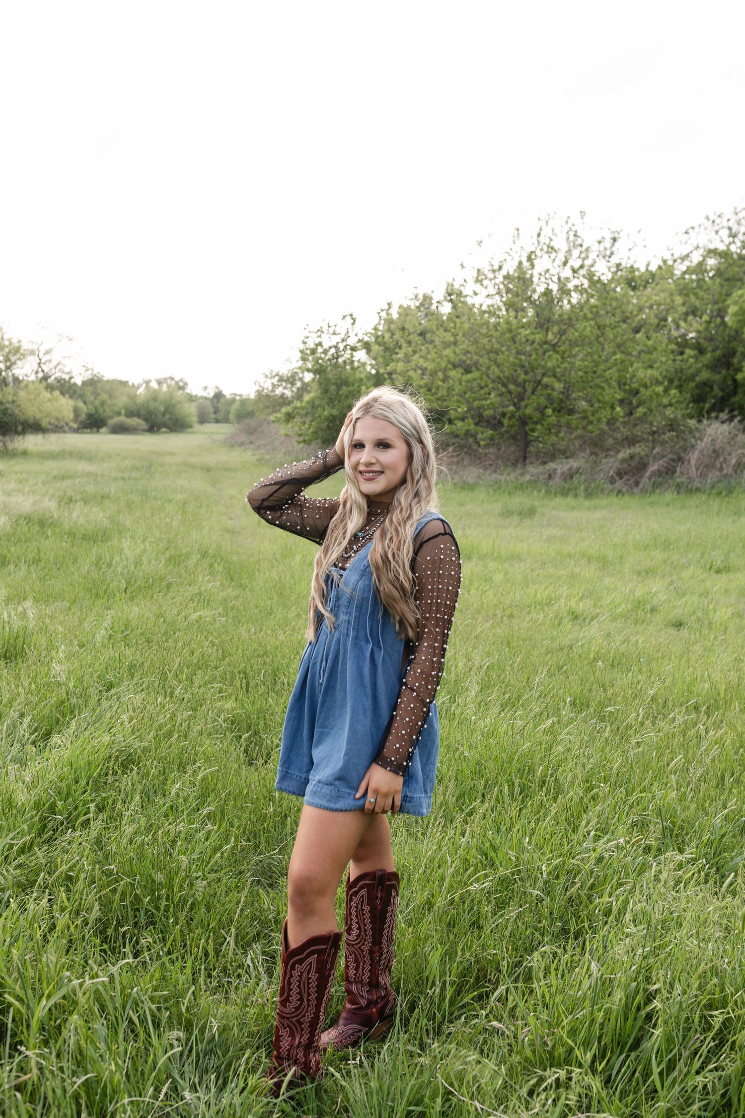 Blonde woman in denim romper, lace top, and cowboy boots posing in a green field.