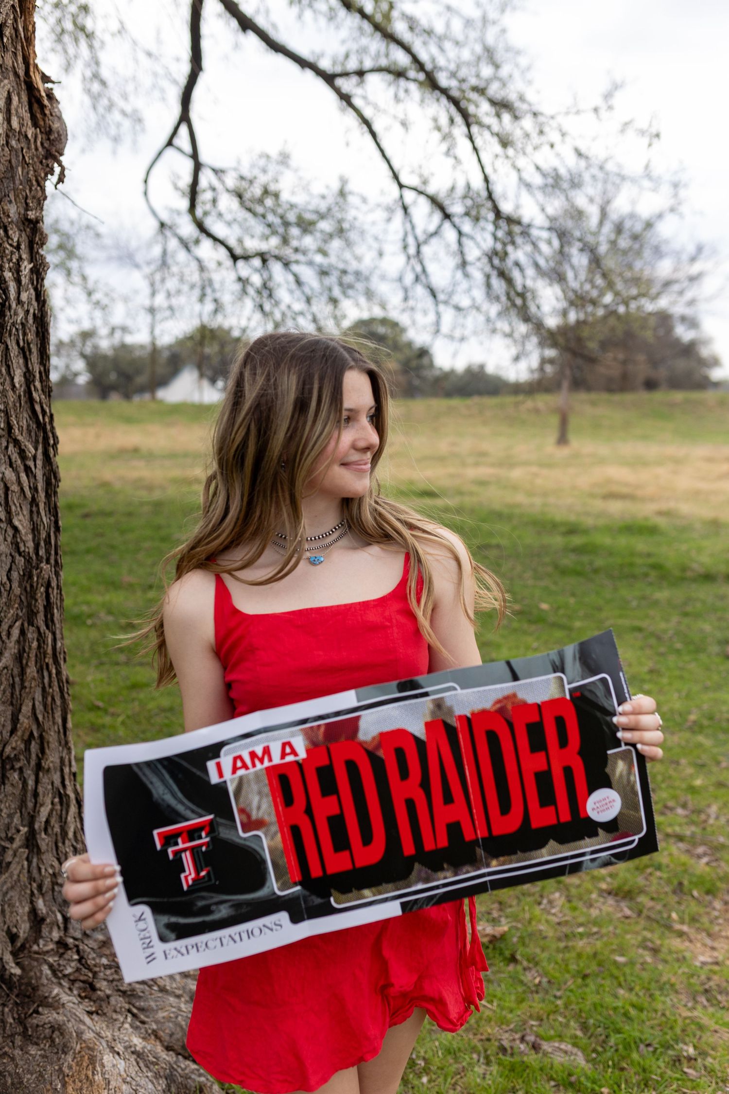 A person in a red dress holds a Texas Tech Red Raiders sign outdoors against a natural backdrop.