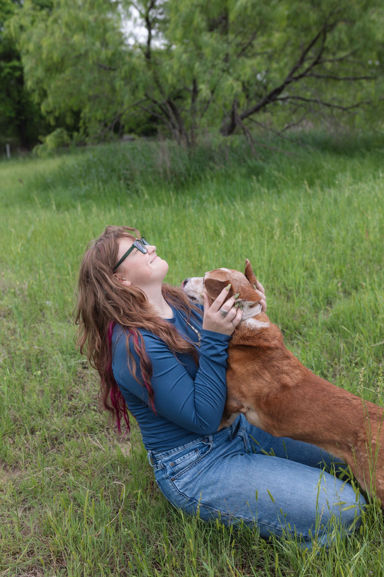 Woman with glasses sitting in green grass field, joyfully holding a brown dog outdoors.