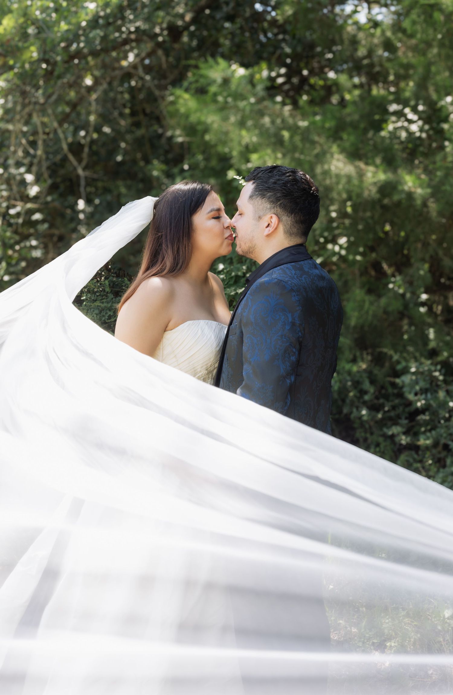 Bride and groom share a romantic kiss outdoors, with a flowing white veil billowing around them against lush green trees.