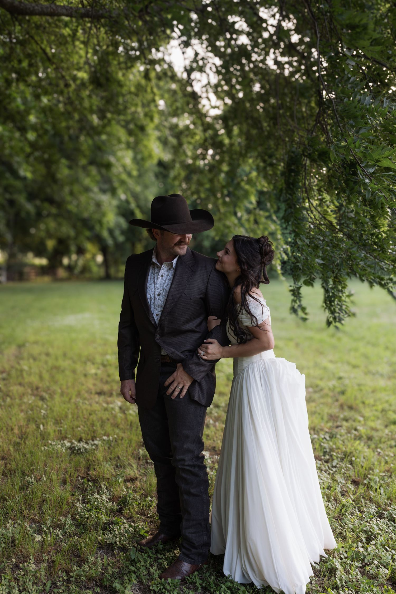 Bride embracing groom in cowboy hat under green tree canopy at rustic outdoor wedding.
