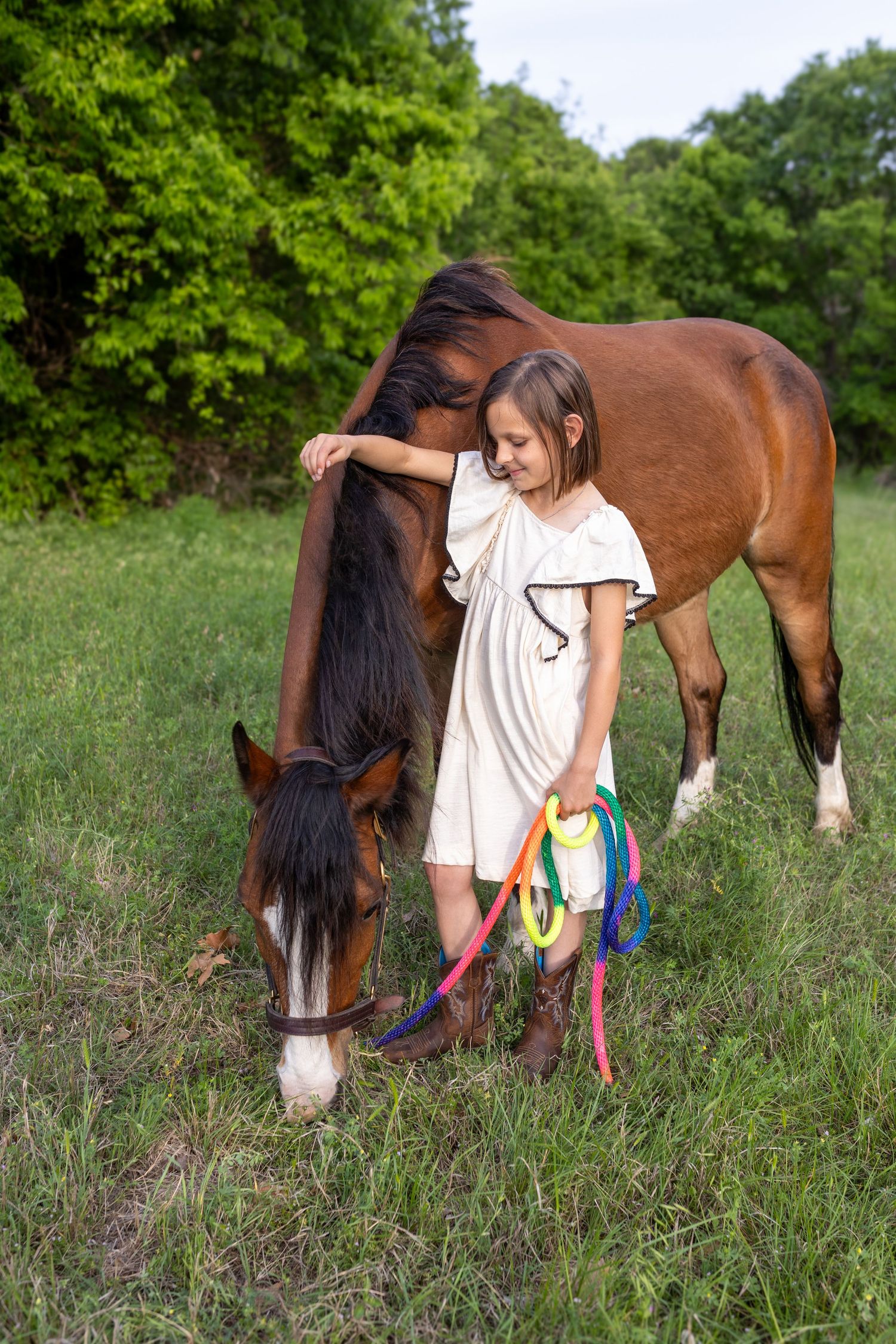Young girl in white dress tenderly embracing a brown horse while holding colorful reins in a green meadow.