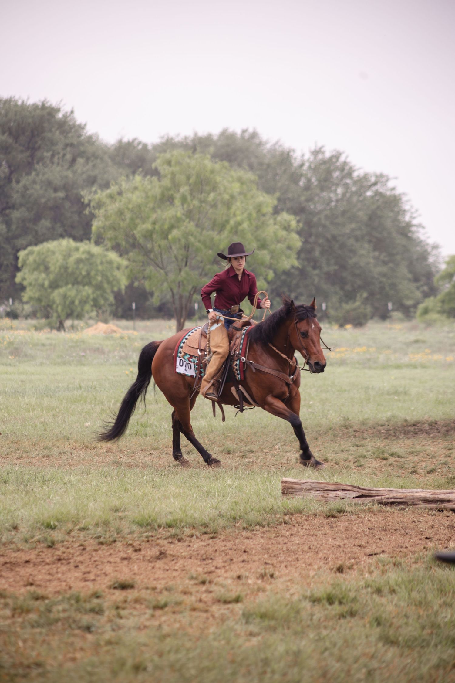 Cowboy in red shirt and hat rides a brown horse across a green field during an outdoor equestrian competition.