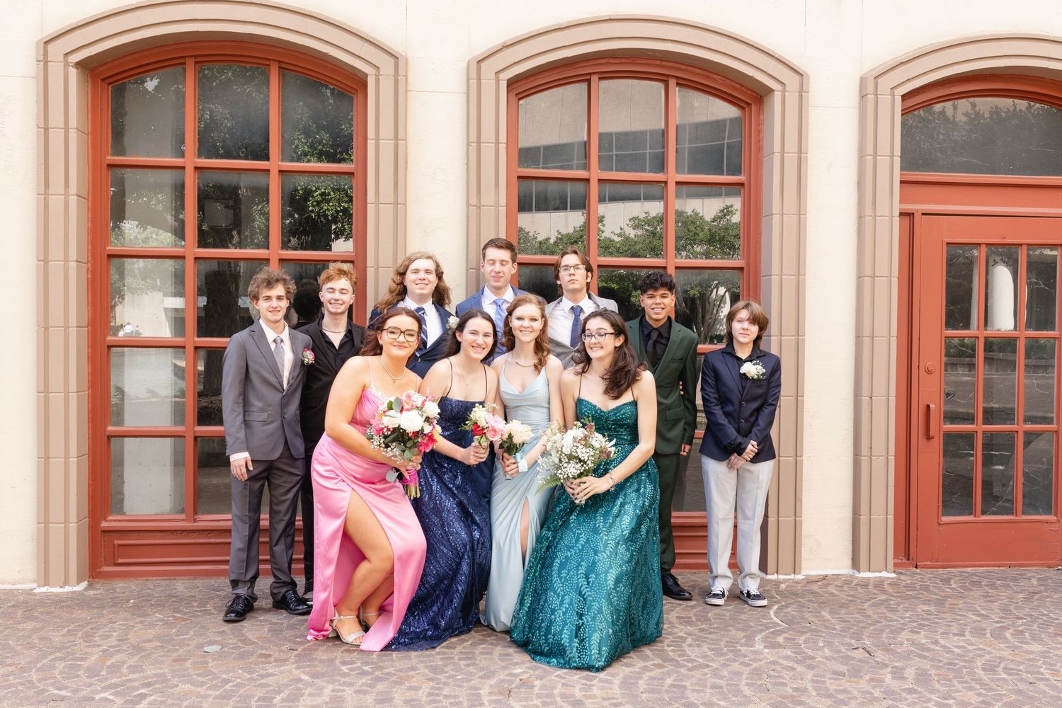 Wedding party group photo with bride, groom, bridesmaids in blue and pink gowns, and groomsmen posing outdoors.