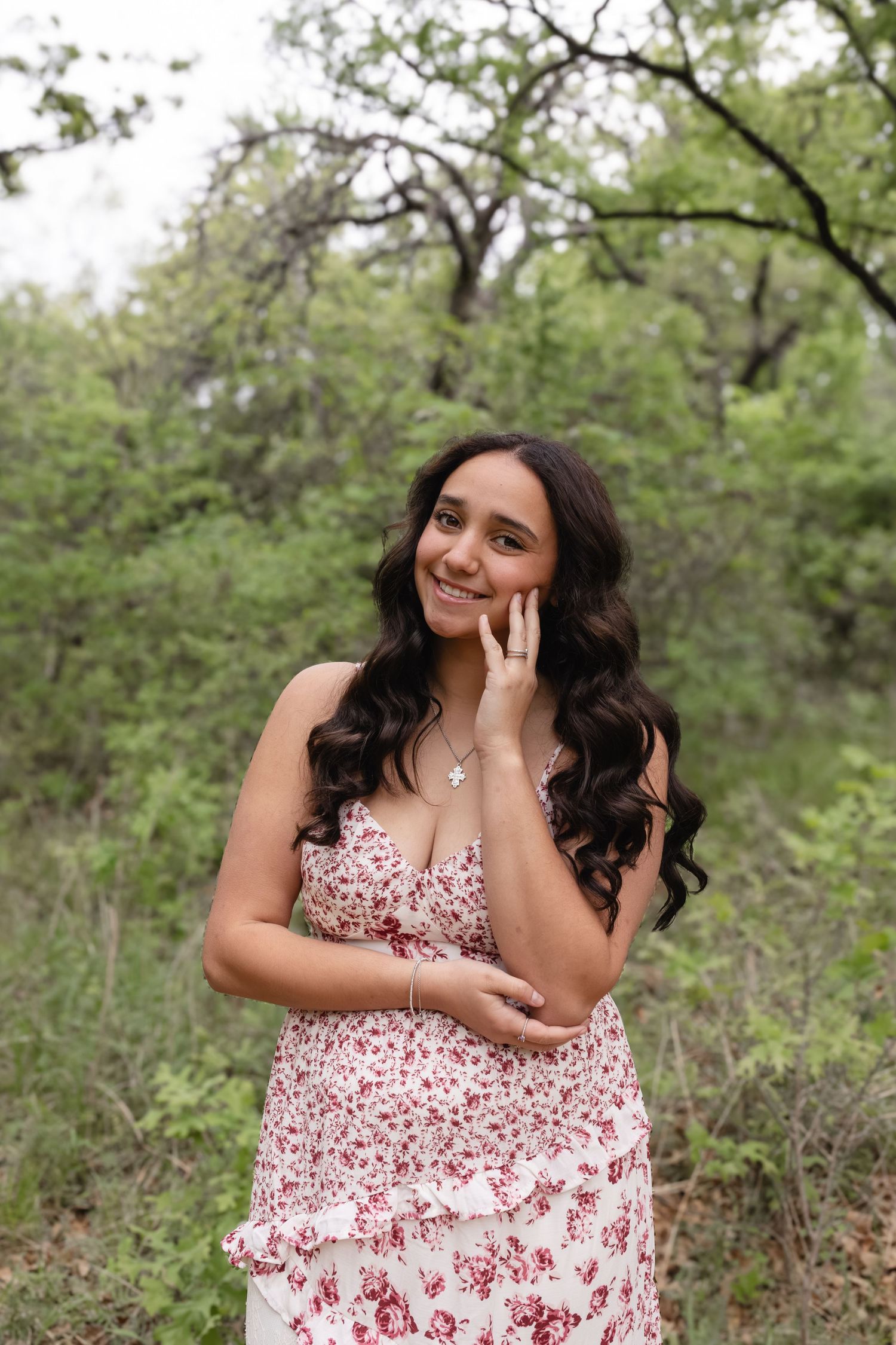Young woman in floral dress poses gracefully outdoors among lush green trees, smiling with hand on cheek.